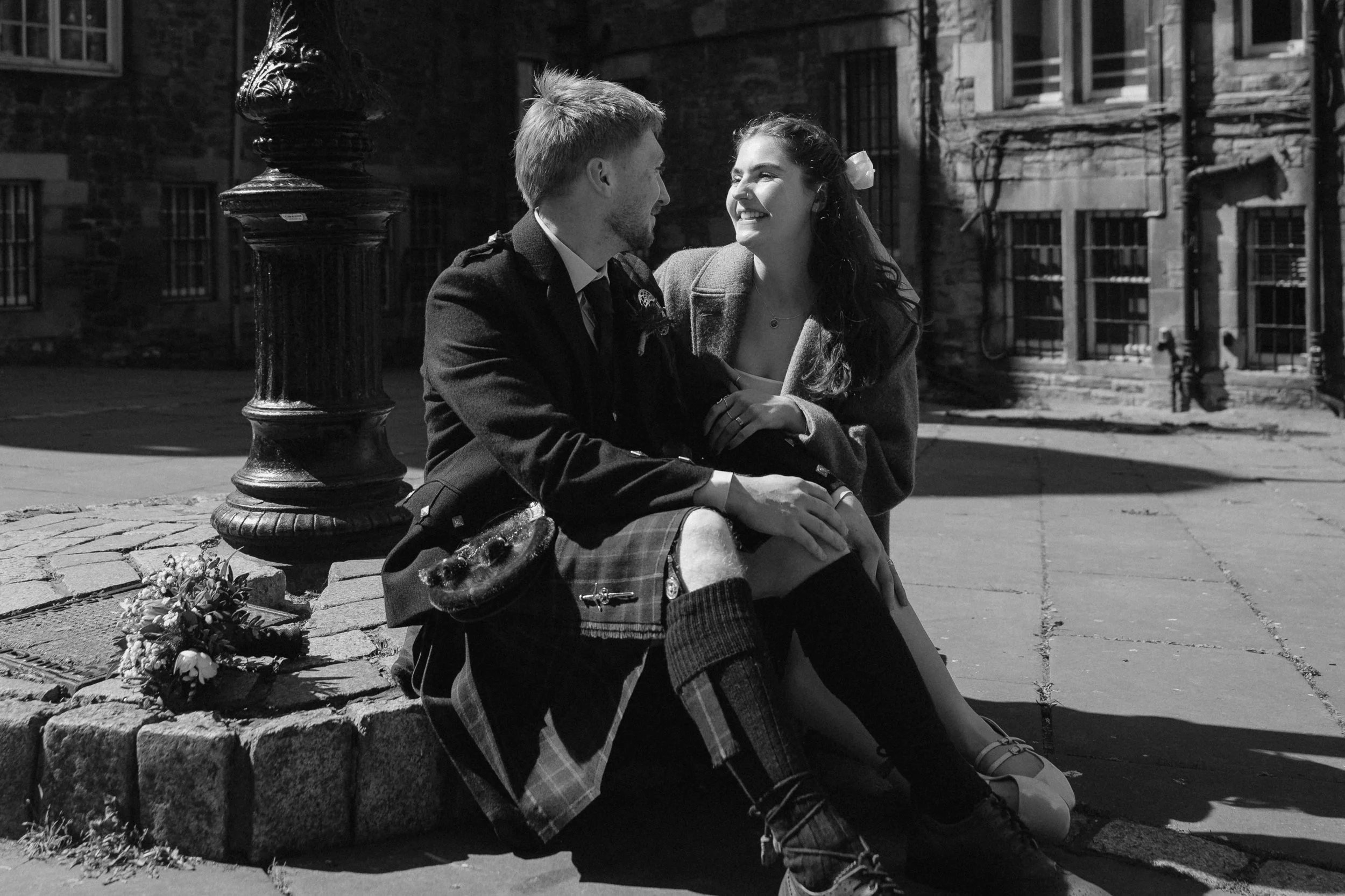 A black-and-white photo of a young couple sitting on a stone platform on a city street, smiling and looking at each other, with a flower bouquet next to them. - captured by an Edinburgh wedding photographer