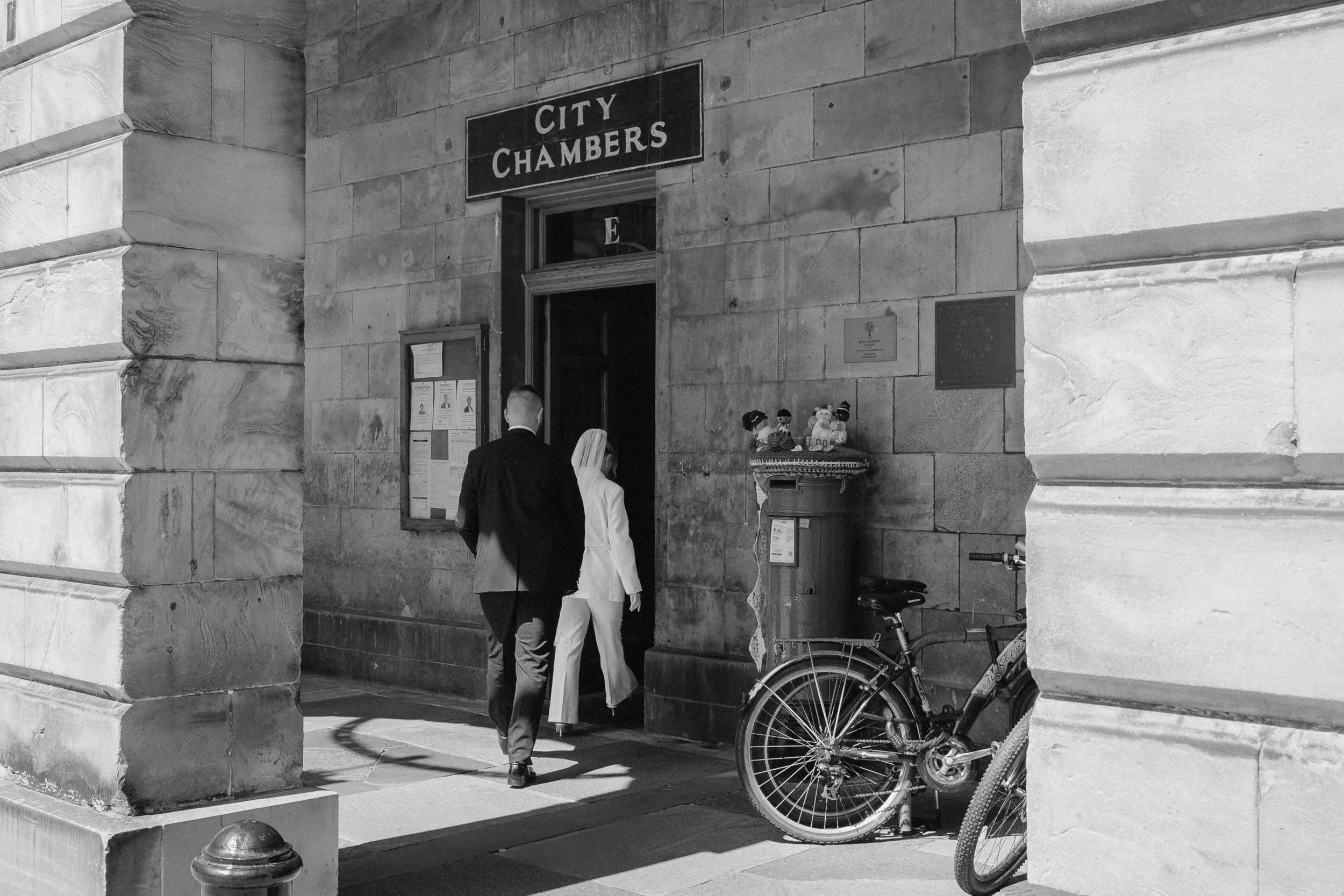 Two people, one in a dark suit and the other in a white suit, walking into a building labeled 'City Chambers.' A bicycle is parked nearby, and there are decorative items on a stand outside. - captured by an Edinburgh wedding photographer