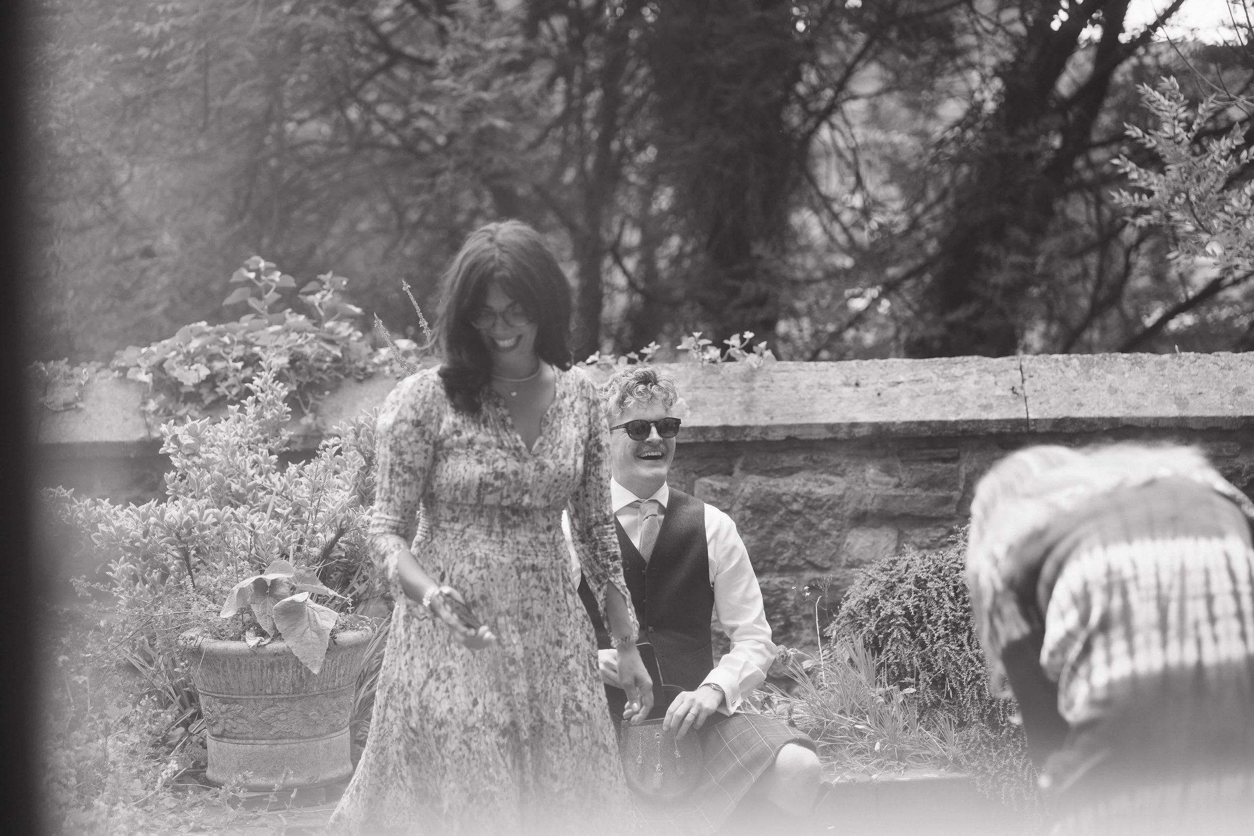 Black and white photo of three people outdoors, a woman in a floral dress and two men, one wearing a kilt and sunglasses, the other in a checked shirt, sitting and standing near a stone wall with trees in the background.