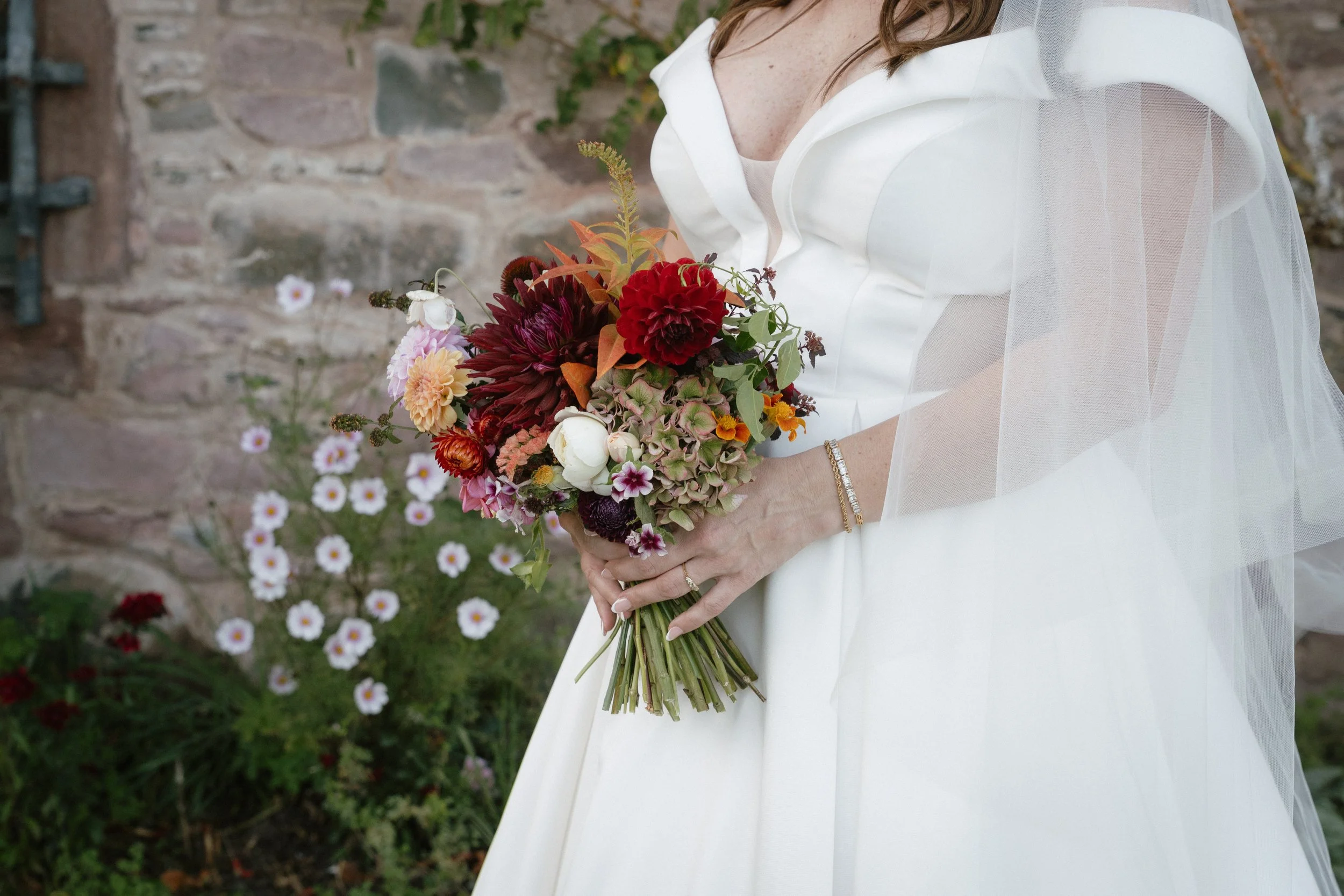 A bride in a white wedding dress holding a bouquet of colorful flowers, standing outdoors near a stone wall and white flowers. - captured by an Edinburgh wedding photographer