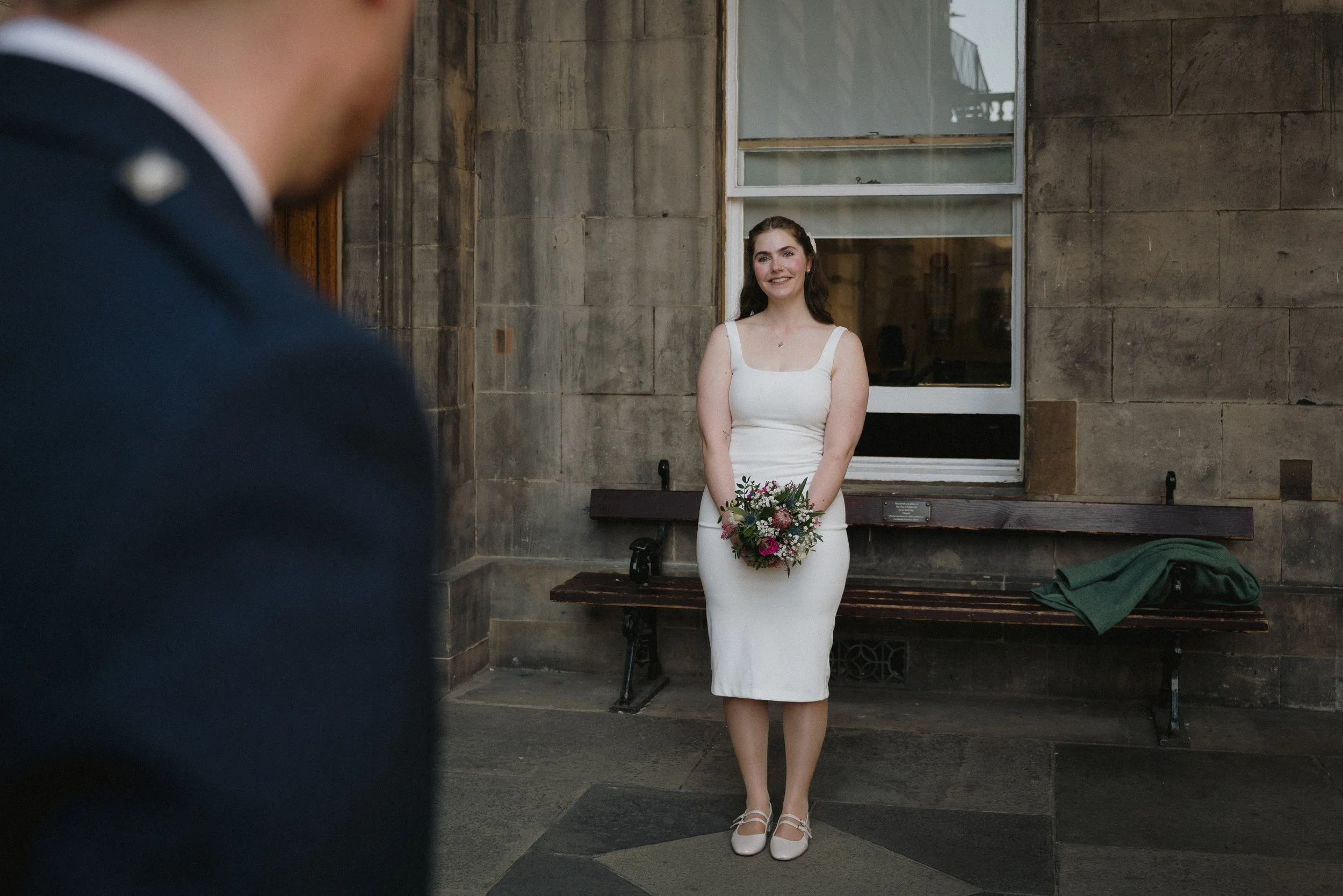 A woman in a white dress holding a bouquet of flowers, standing in front of a stone building and looking at a person in a suit who is partially visible in the foreground. - captured by an Edinburgh wedding photographer