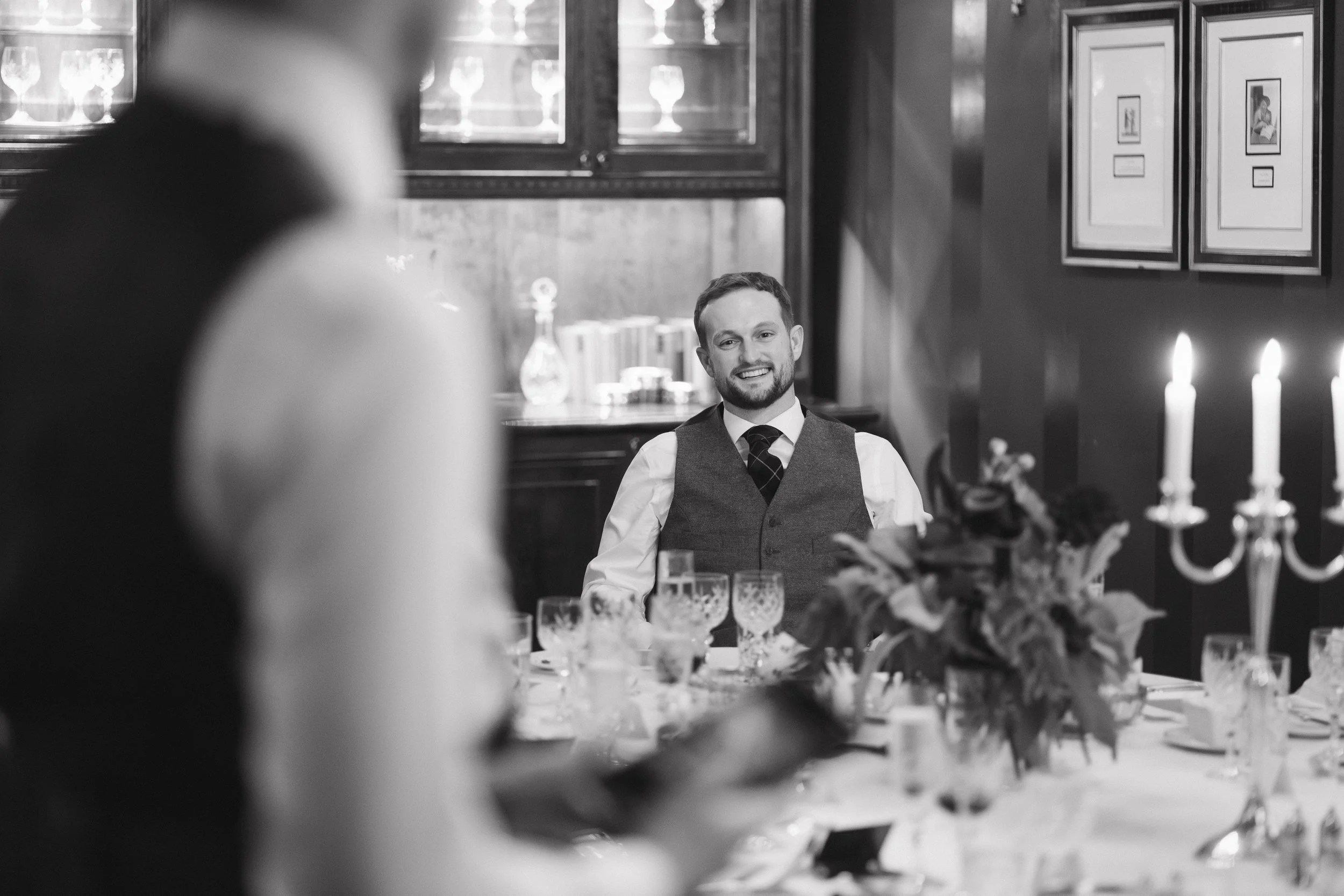 A smiling man in formal attire sitting at a decorated table, with blurred foreground of a woman, in a dimly lit room with framed pictures on the wall and lit candles. - captured by an Edinburgh wedding photographer
