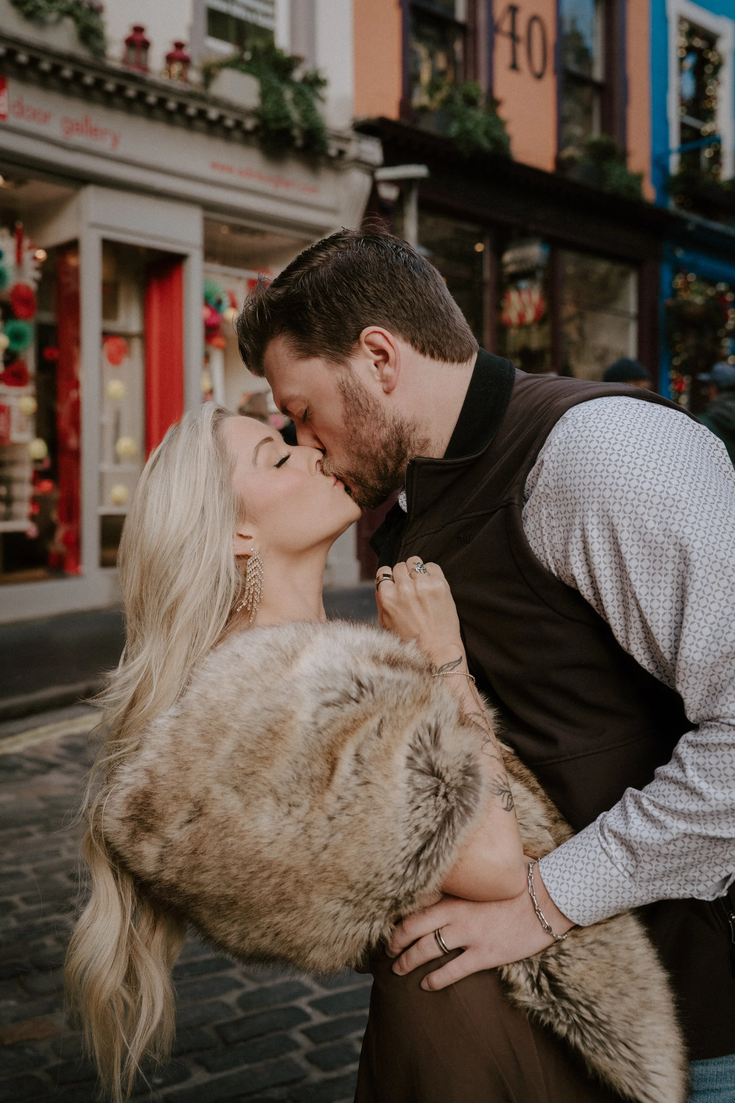 A couple kissing on a city street during the holidays, with the woman holding a large fur coat. - captured by an Edinburgh wedding photographer