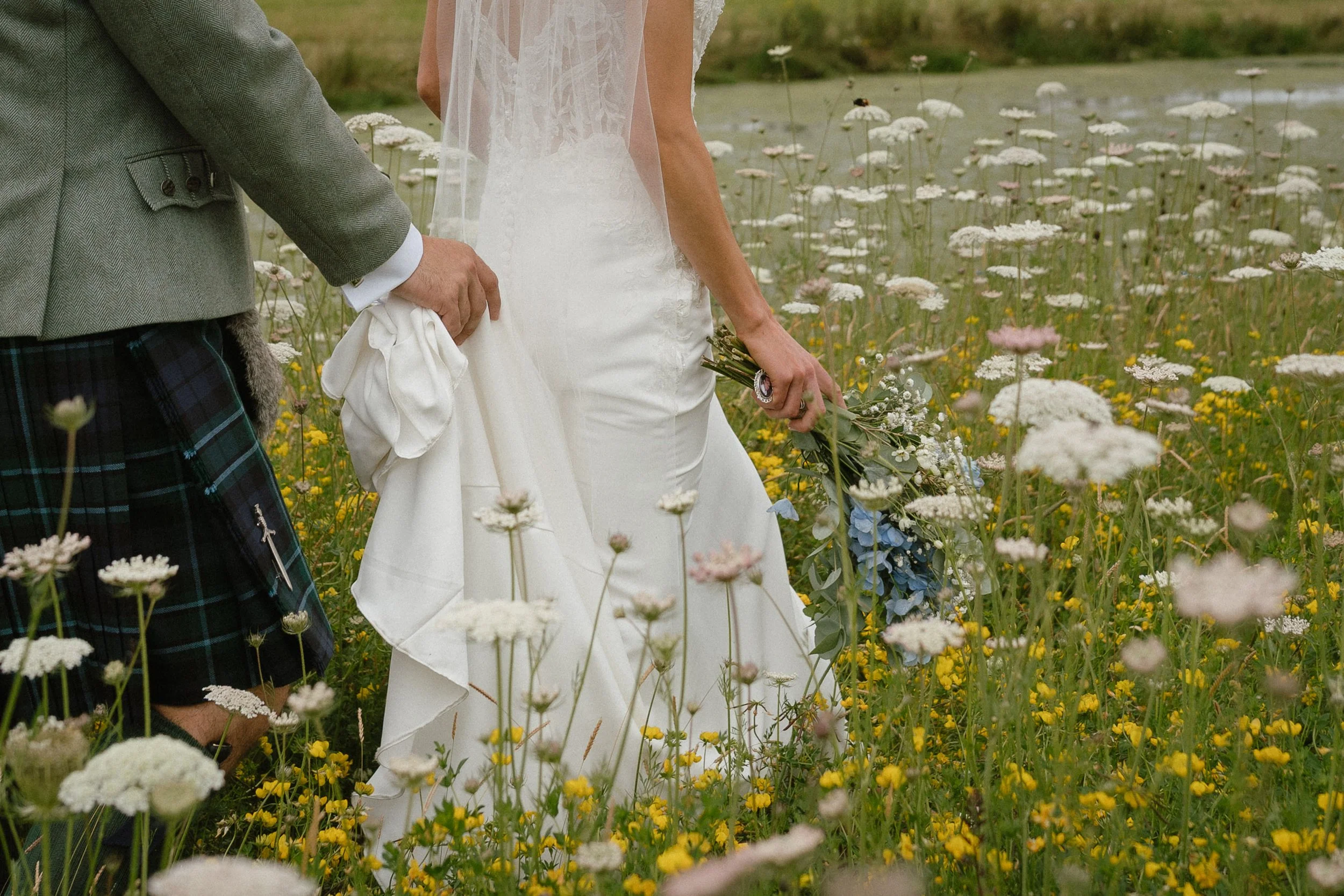 A bride holding a bouquet of flowers and a groom dressed in traditional Scottish attire, standing in a field of wildflowers. - captured by an Edinburgh wedding photographer