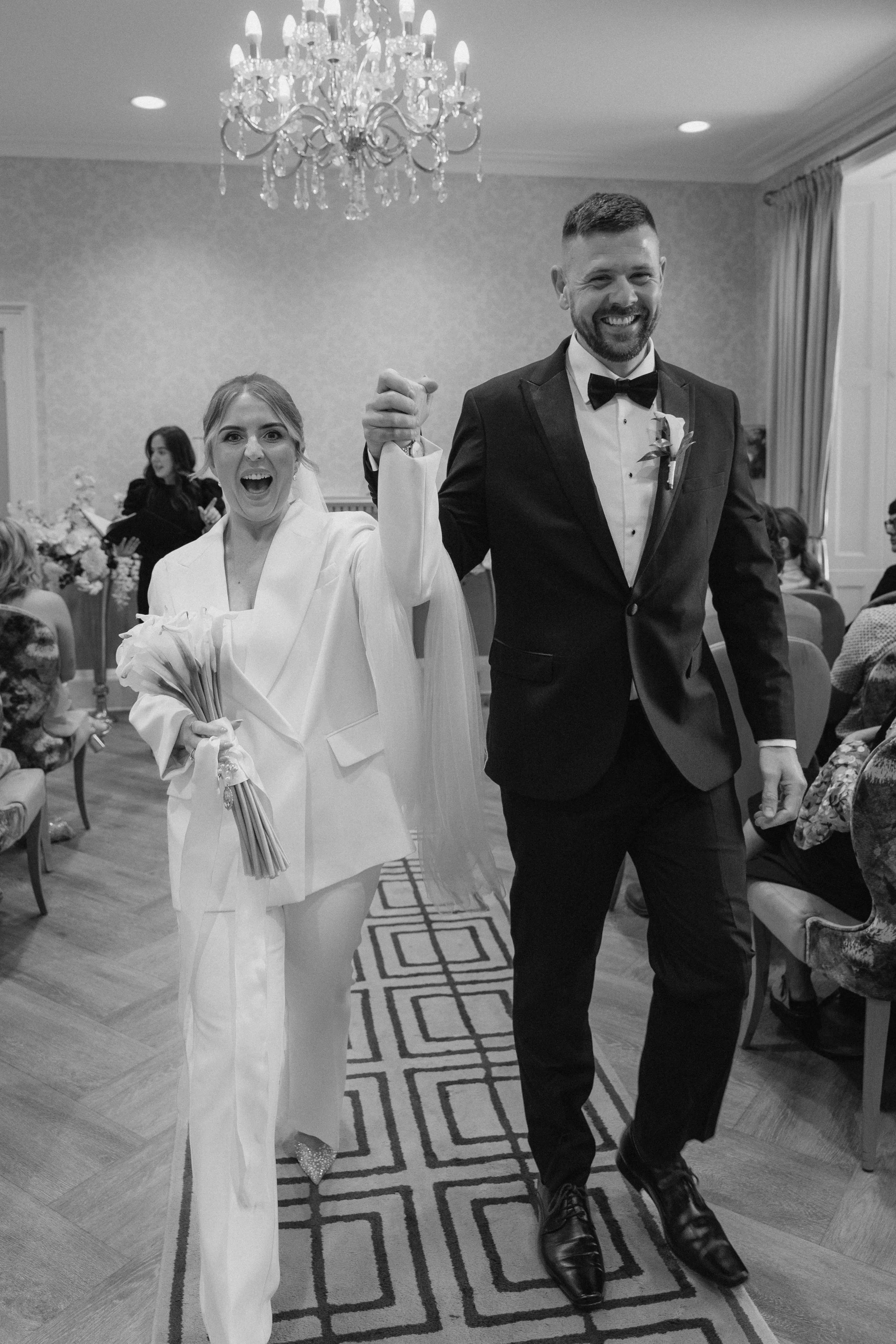 A black-and-white photo of a couple walking down the aisle at their wedding reception, holding hands and smiling. The woman is wearing a white coat and holding a bouquet, while the man is dressed in a black tuxedo.