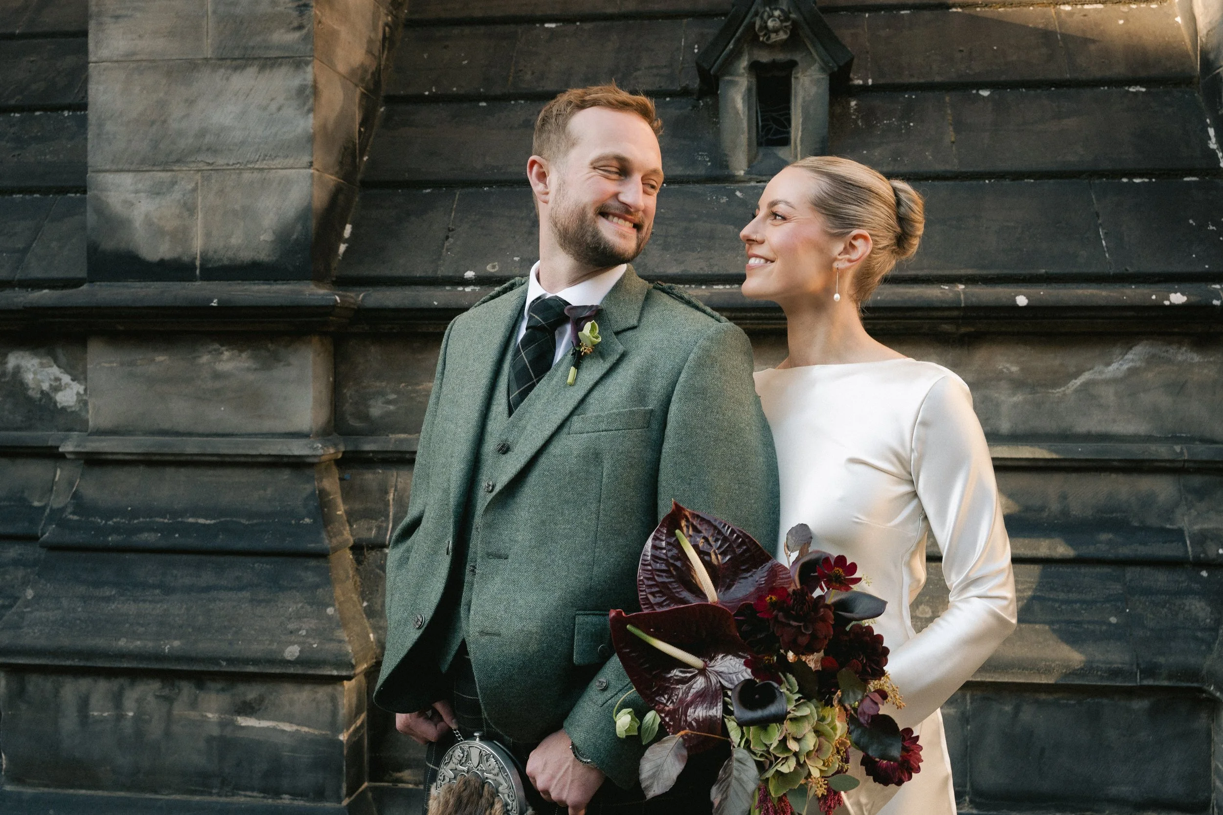 A newlywed couple stands close to each other on a stone building exterior. The man is dressed in a gray suit with a green tartan tie and a boutonniere. The woman wears a white satin wedding dress and holds a dark red and green floral bouquet. They ar