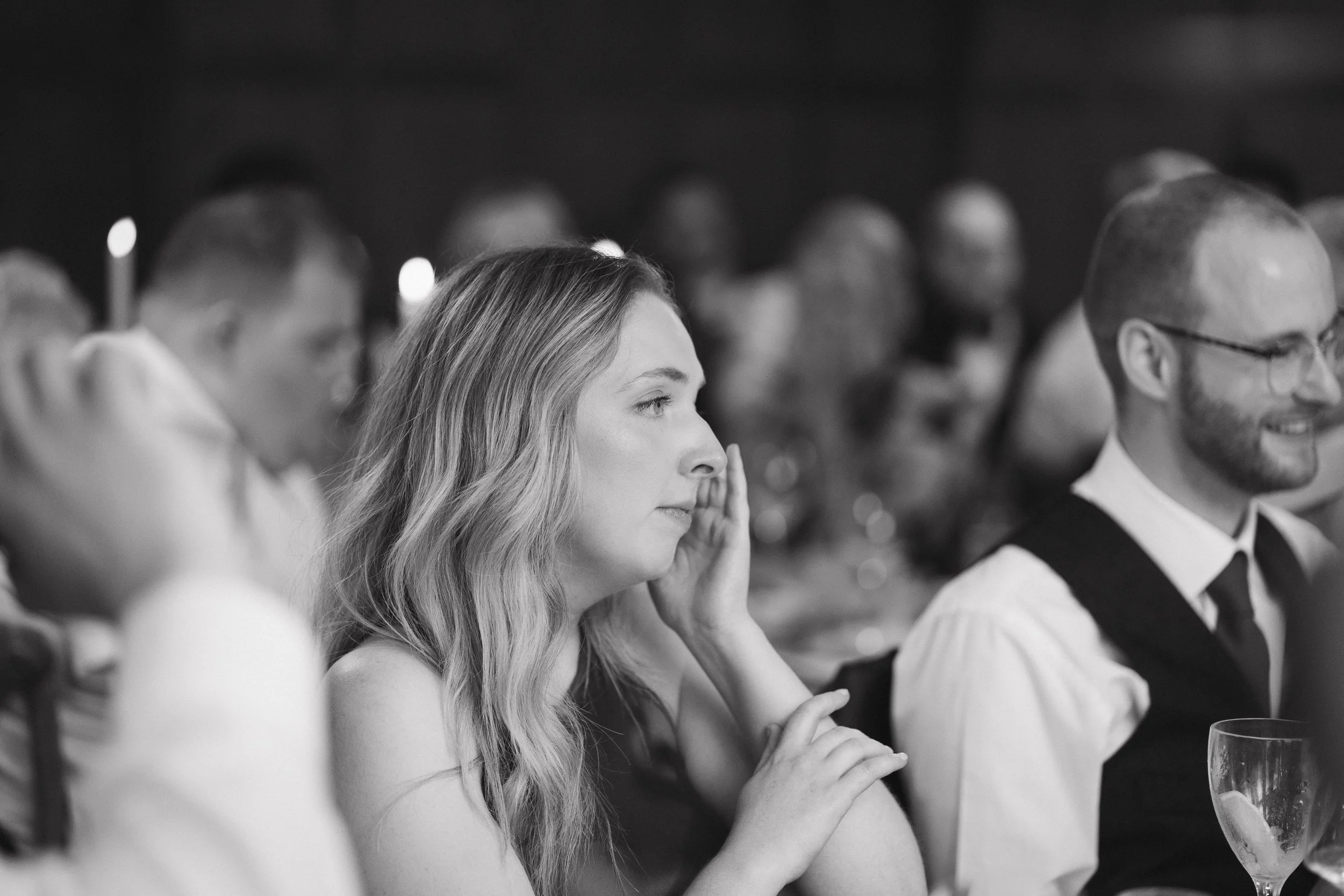 Black and white photo of a woman with long wavy hair attentively listening at a formal event, with a man with glasses and a beard sitting next to her smiling, surrounded by other seated guests. - captured by an Edinburgh wedding photographer