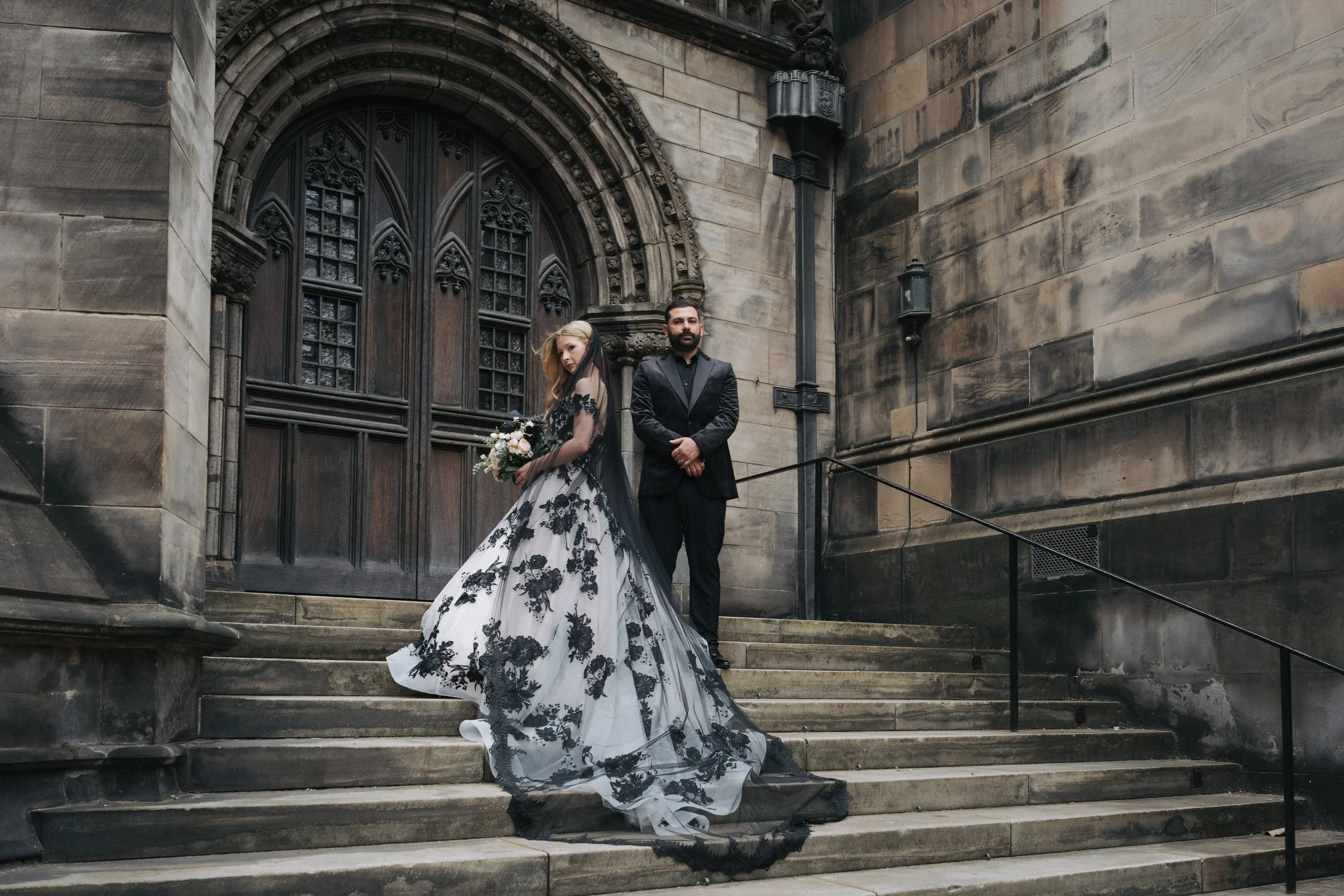 A bride in a black and white wedding gown holding a bouquet stands on stone steps beside a groom in a black tuxedo, in front of a large wooden door of a historic stone building. - captured by an Edinburgh wedding photographer
