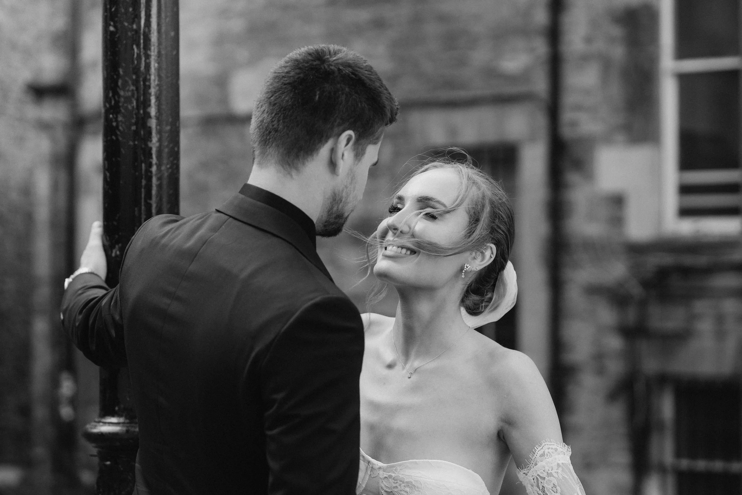 A couple smiling and looking at each other outdoors, with the woman touching her hair and wearing a lace dress, and the man dressed in a suit. Black and white photo. - captured by an Edinburgh wedding photographer