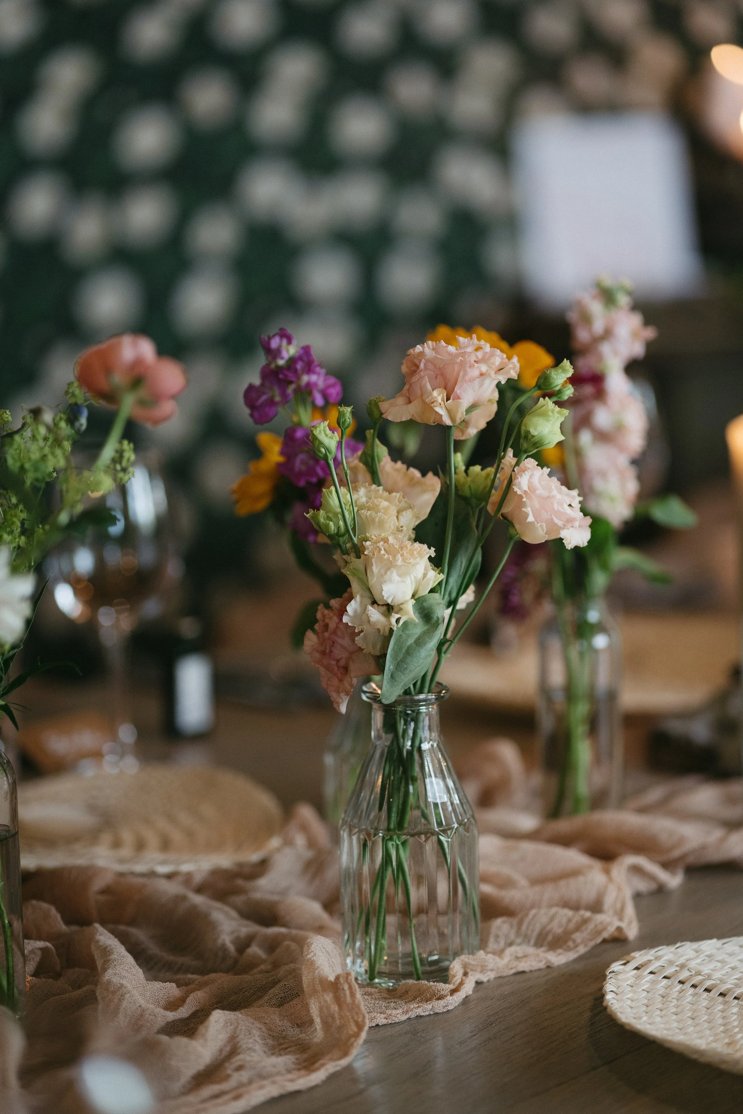 A table centerpiece with a glass bottle containing a bouquet of colorful flowers, set on a rustic table with a soft cloth, in a dimly lit room. - captured by an Edinburgh wedding photographer