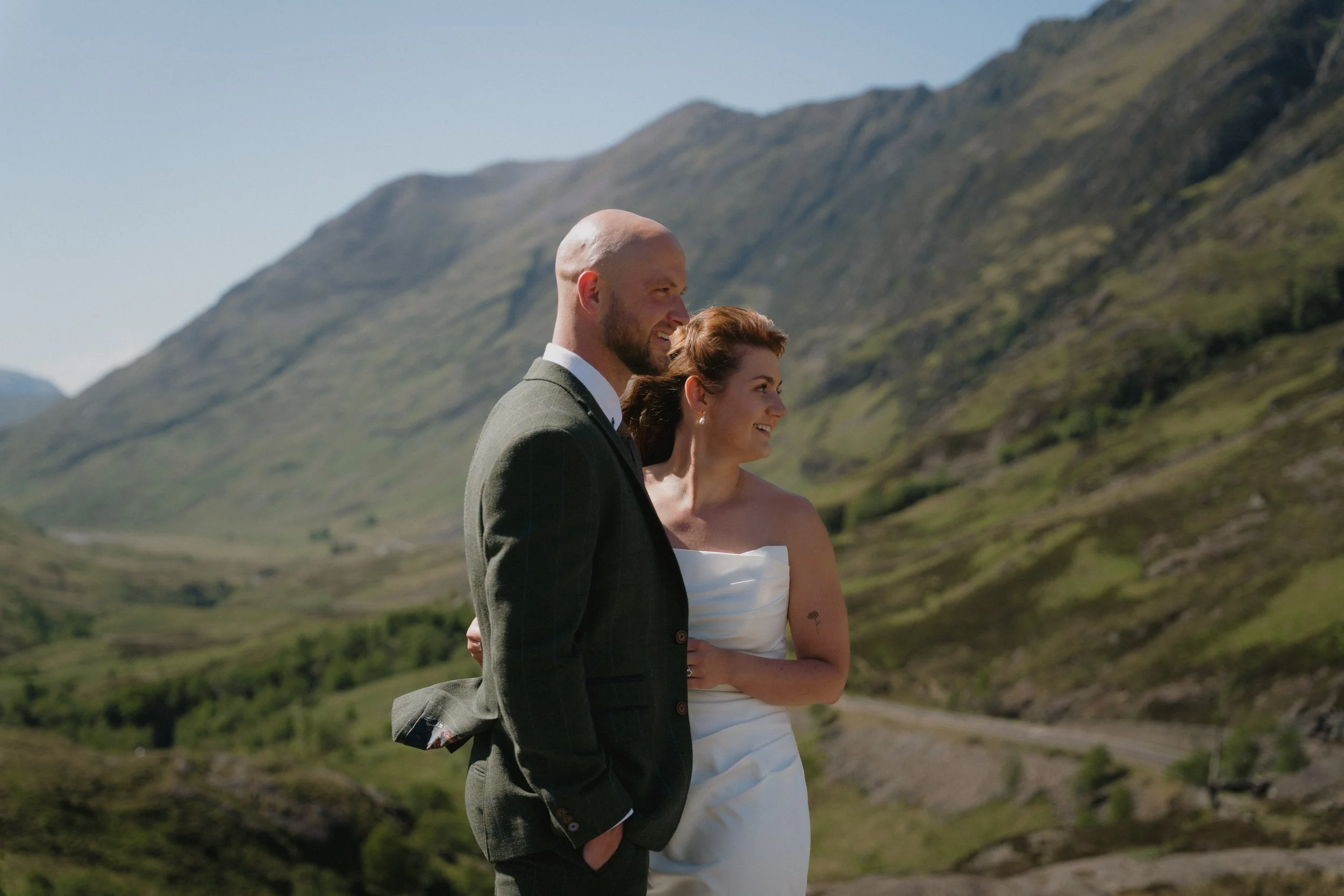 A recently married couple standing outdoors in a scenic mountainous area, smiling and enjoying the moment.