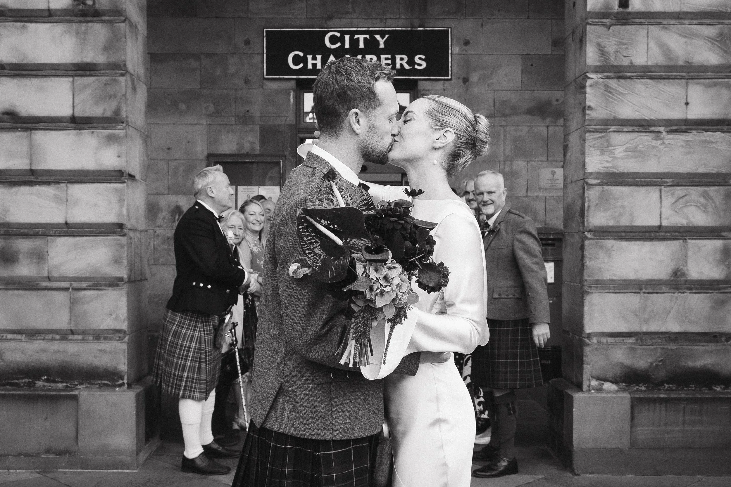 A black and white photo of a newlywed couple sharing a kiss, with the bride holding a bouquet, in front of a brick city hall entrance. Several onlookers dressed in traditional Scottish attire smile in the background.