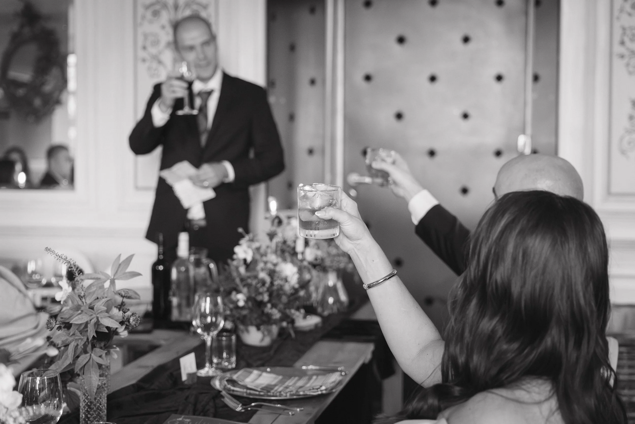 People raising glasses in a toast at a formal gathering or celebration, with a man in a suit in the background holding a glass and smiling. - captured by an Edinburgh wedding photographer