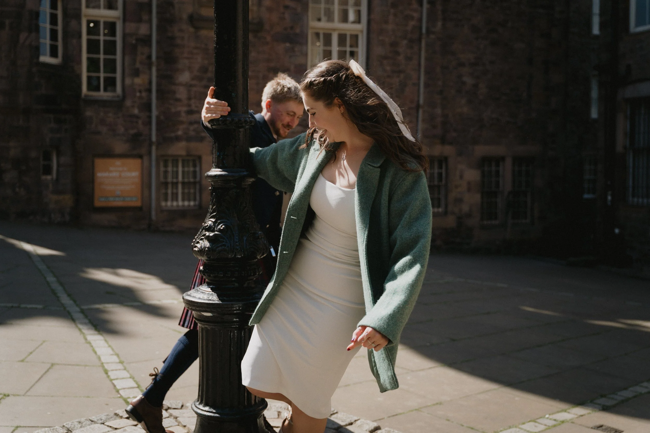 A woman in a white dress and green coat laughs and leans against a black lamppost, while a man in the background smiles behind her. - captured by an Edinburgh wedding photographer