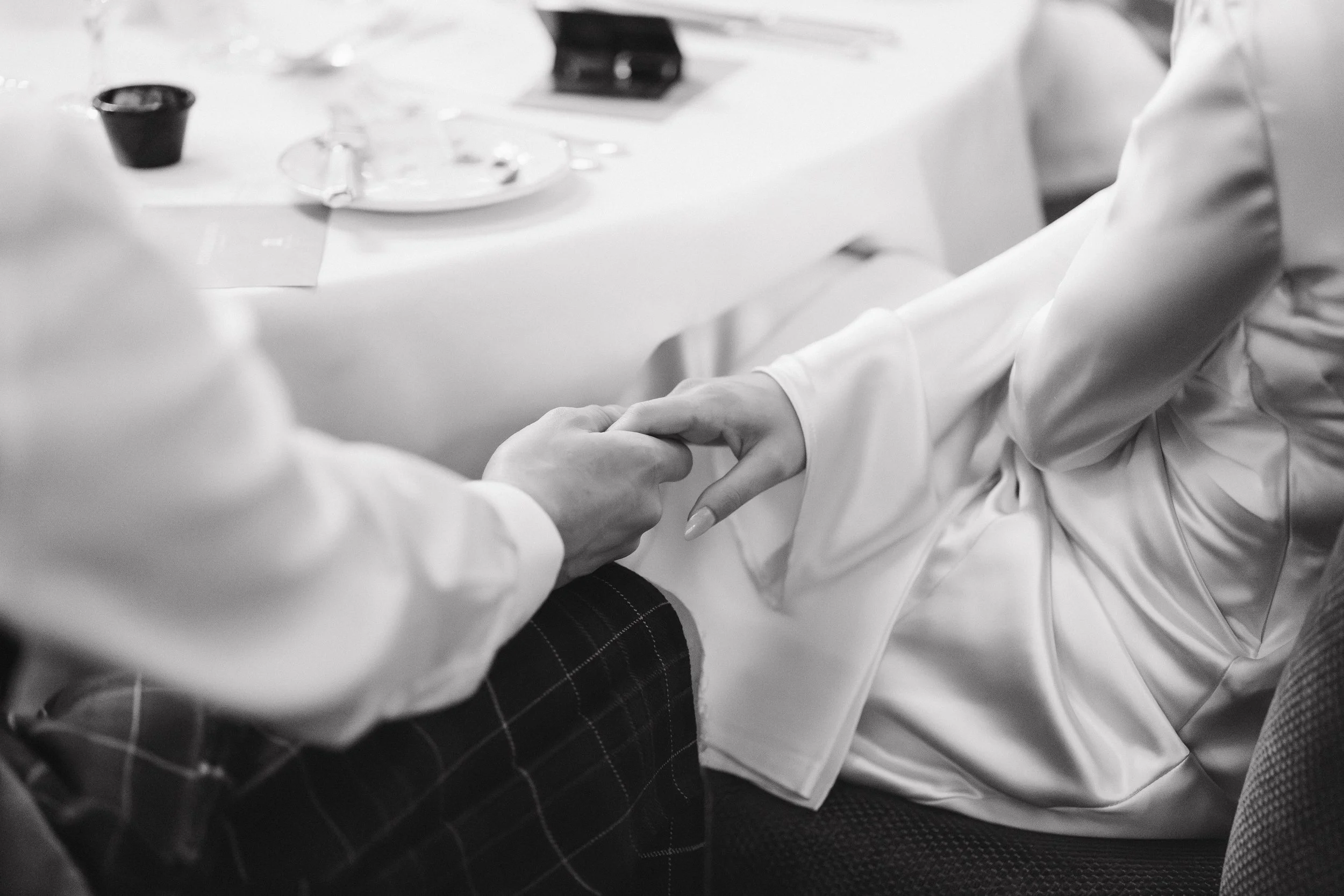 A black-and-white photo of two people holding hands across a dinner table, with tableware and a black cup visible in the background. - captured by an Edinburgh wedding photographer