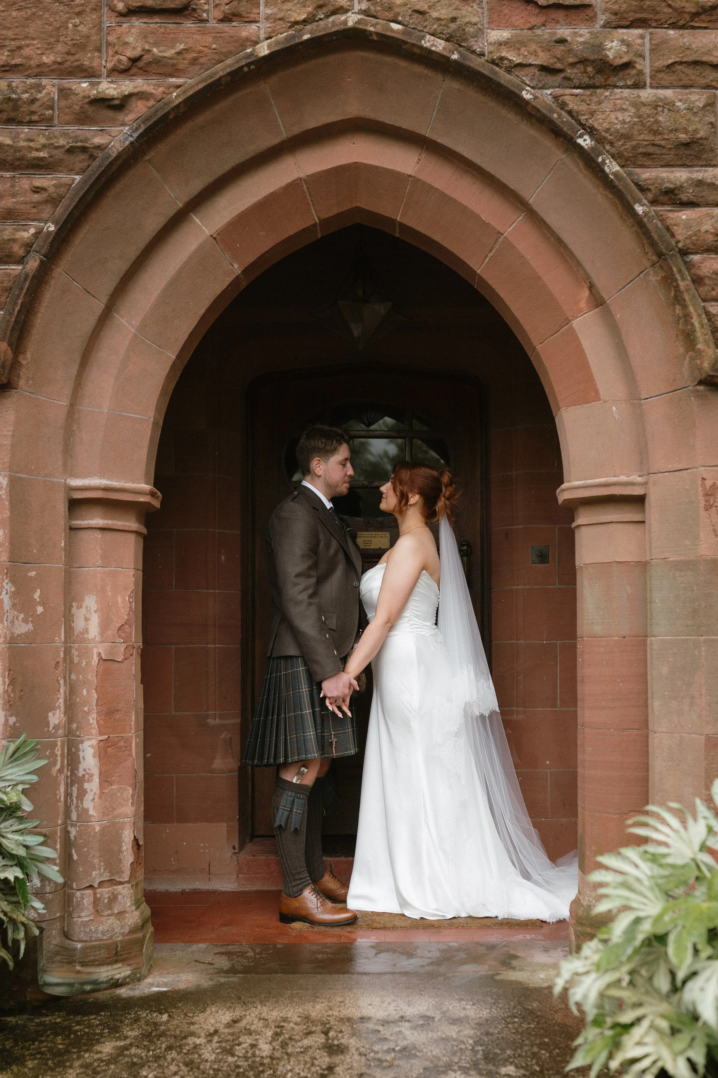 A bride and groom holding hands in front of a brick archway during their wedding ceremony. - captured by an Edinburgh wedding photographer