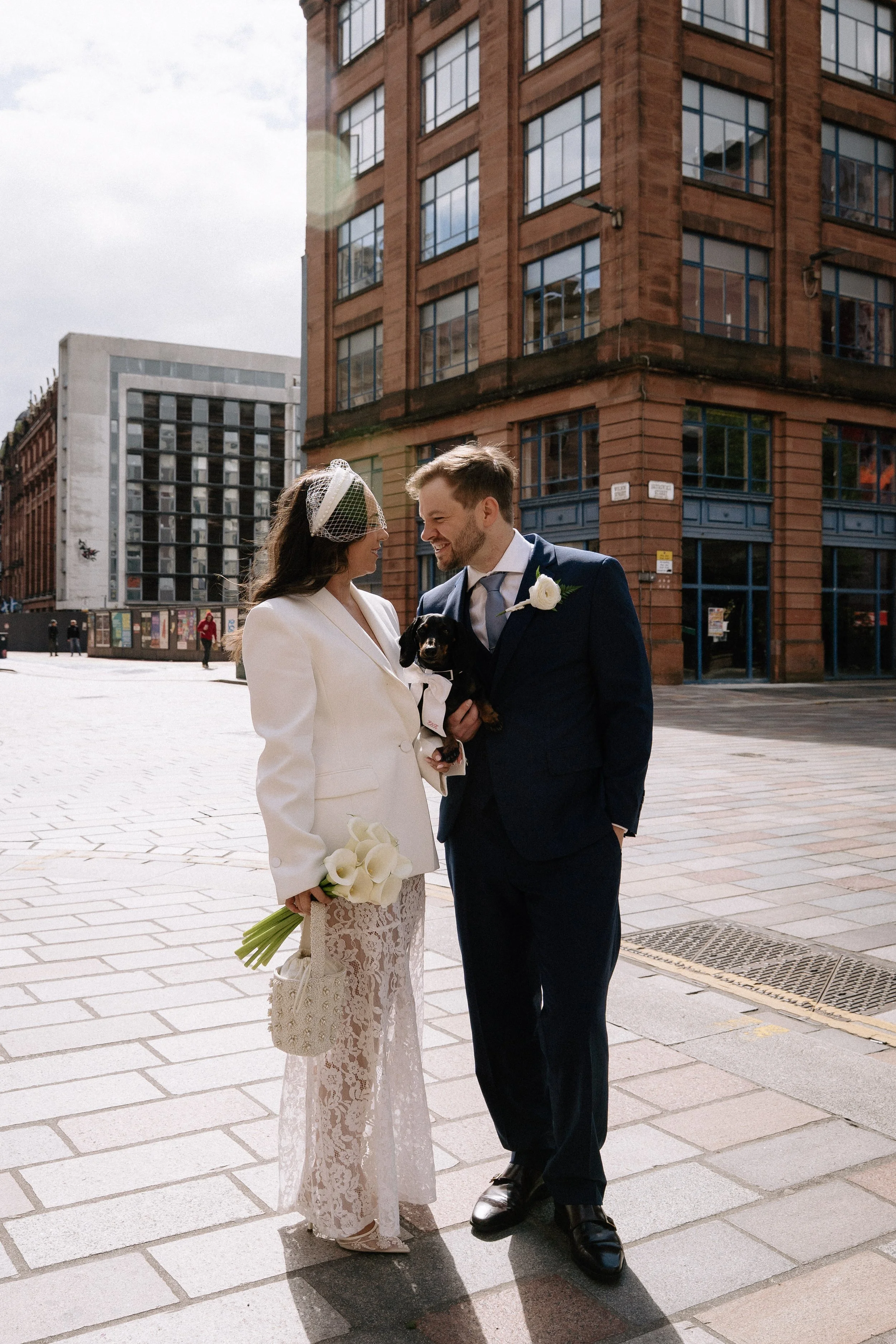 A wedding couple smiling and looking at each other on a city street, with the bride holding a bouquet of white calla lilies and the groom holding a small black puppy. - captured by an Edinburgh wedding photographer