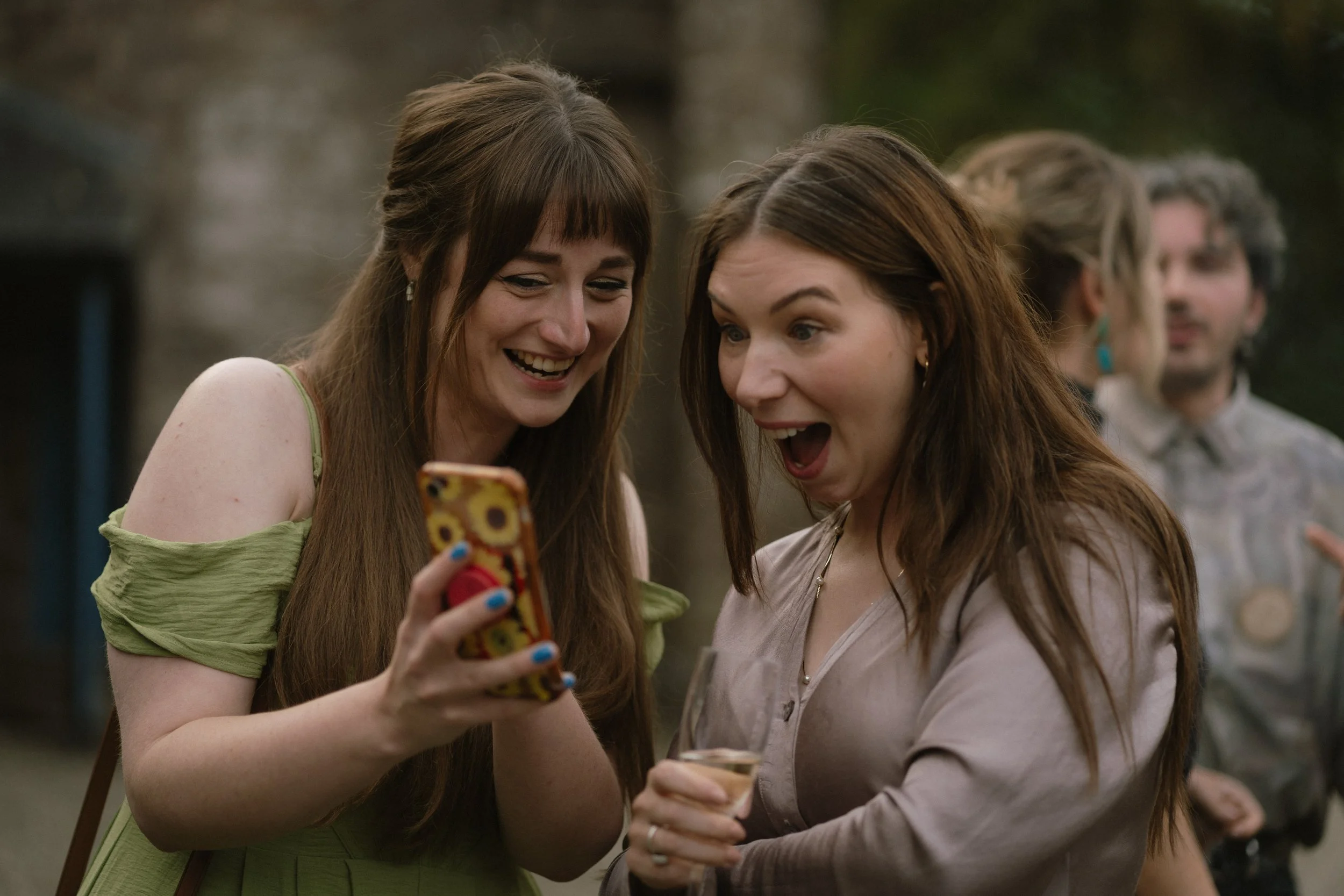 Two women are looking at a smartphone with excitement and happiness, one holding a glass of wine, in an outdoor setting with blurred people in the background. - captured by an Edinburgh wedding photographer