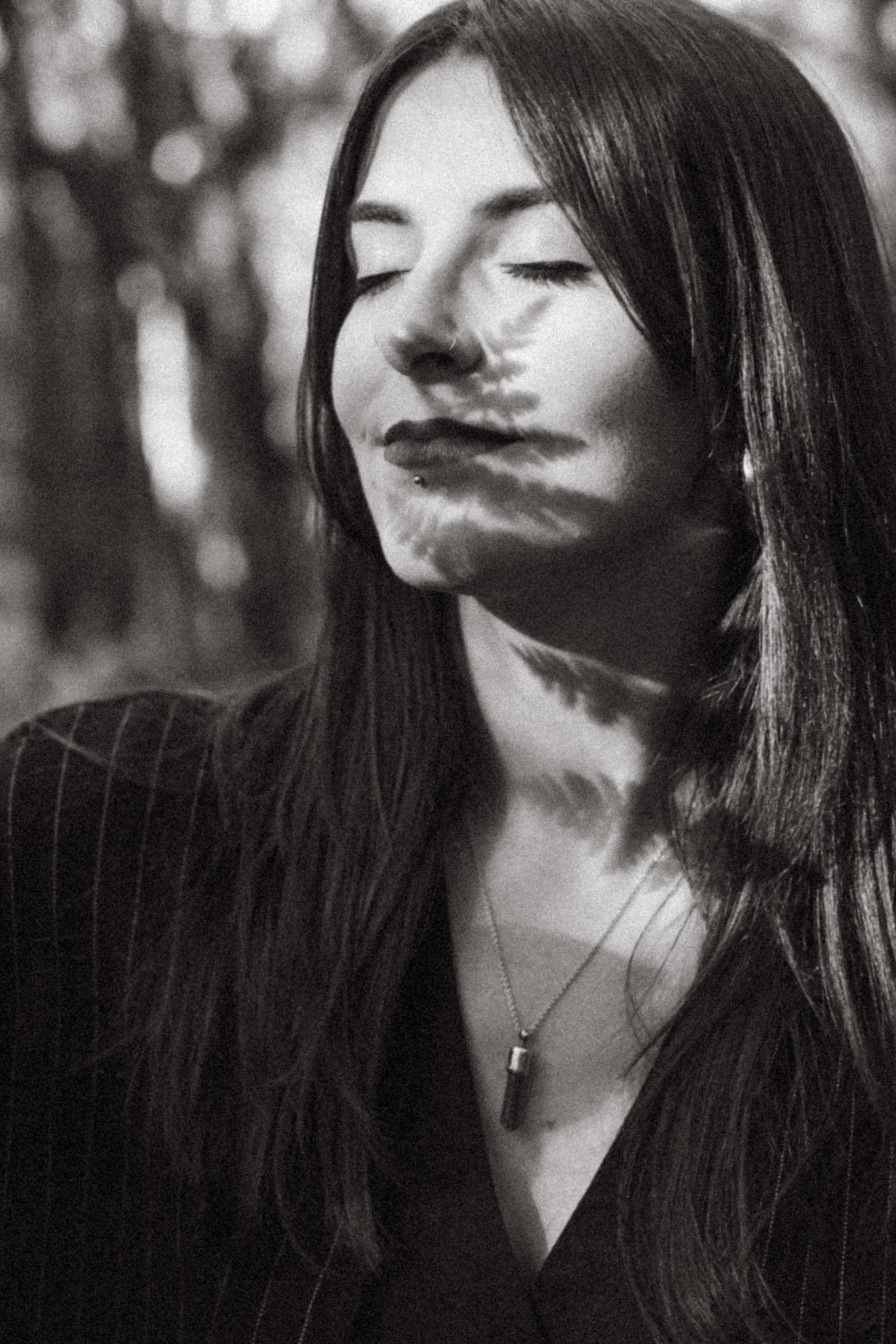 Black and white photo of a woman with long, straight hair, eyes closed, and a subtle smile. She is wearing a pinstripe blazer and a necklace with a pendant.