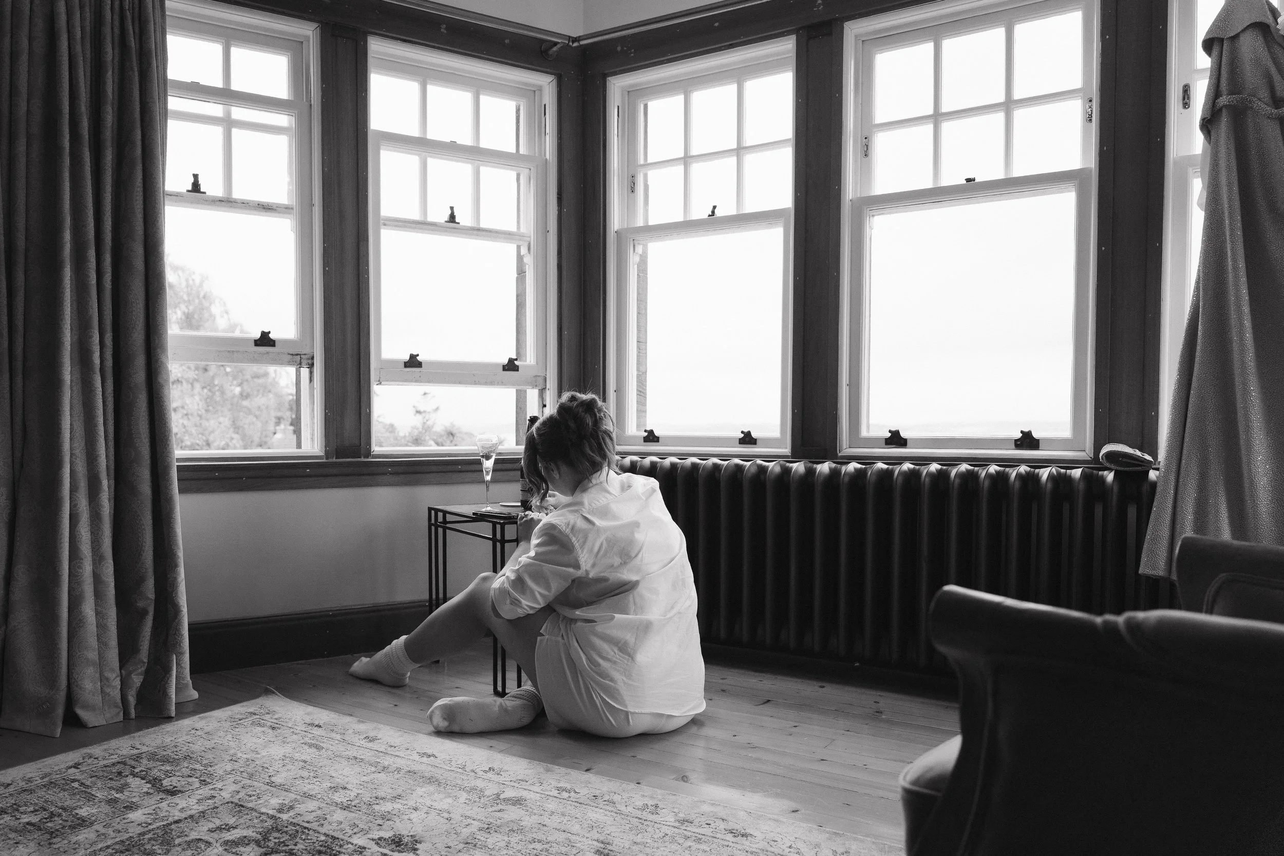 A woman sits on the floor by a window, wearing a white shirt and socks, with a glass of champagne on a small table next to her. - captured by an Edinburgh wedding photographer
