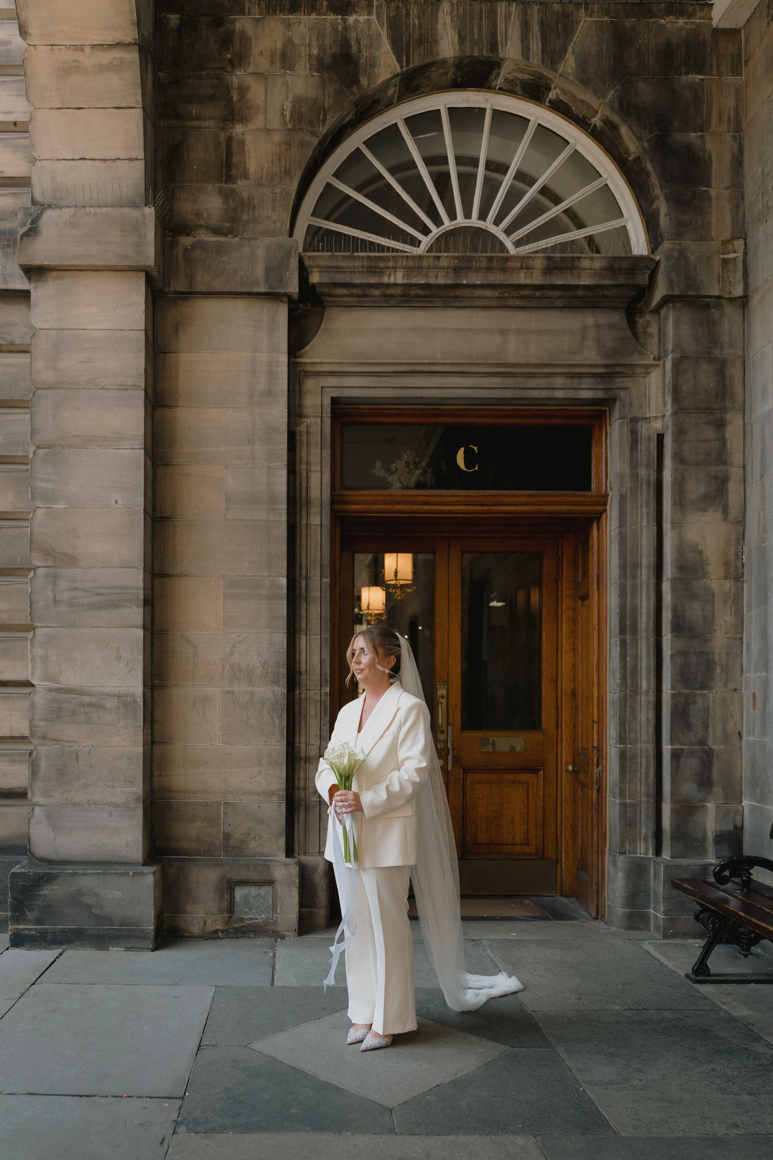 Bride in a white suit holding a bouquet of white calla lilies standing outside a historic stone building with a large wooden door and a semi-circular window above. - captured by an Edinburgh wedding photographer