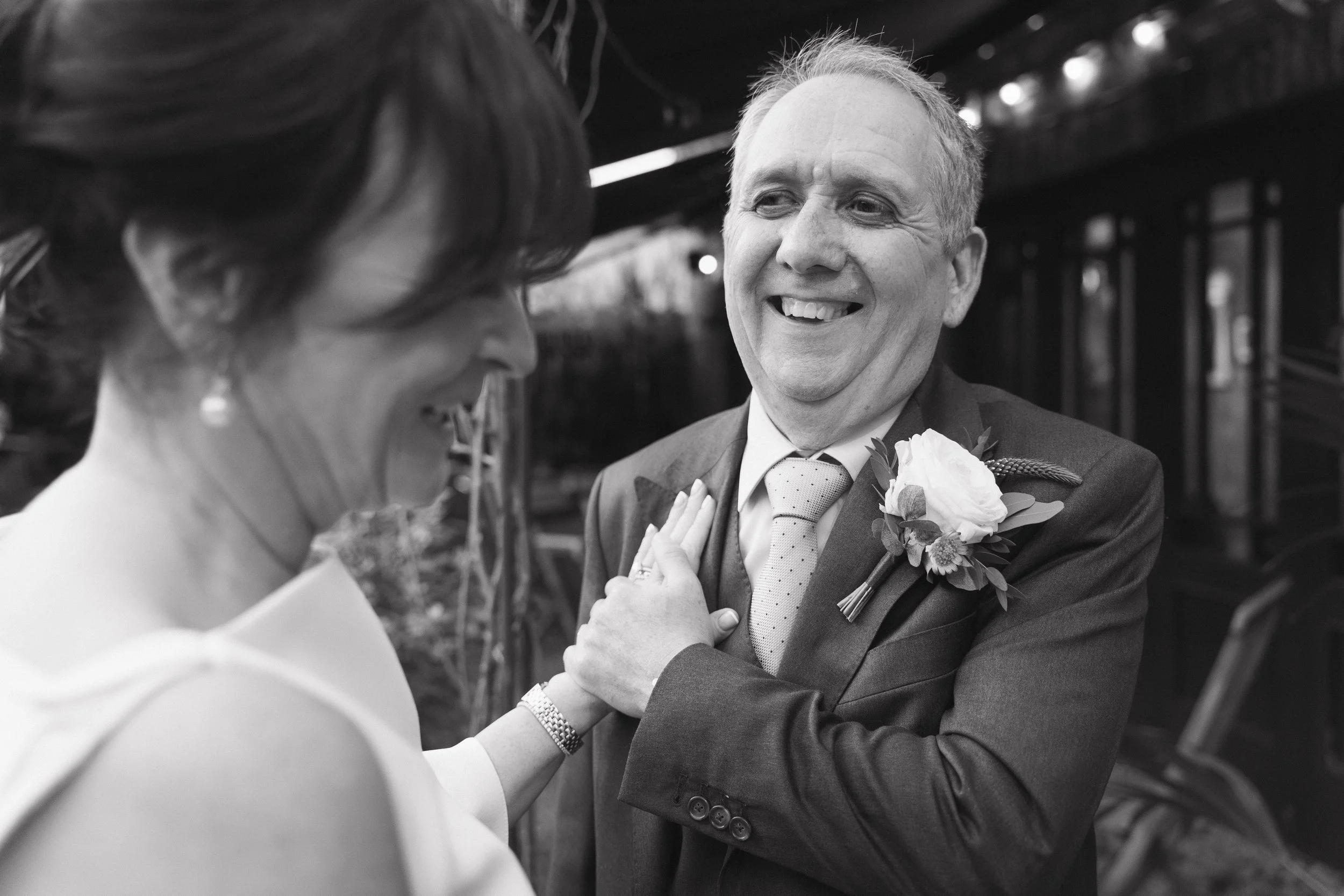 A joyful man in a suit with a boutonniere on his lapel smiling at a woman, who is smiling back, in a black and white photo. - captured by an Edinburgh wedding photographer