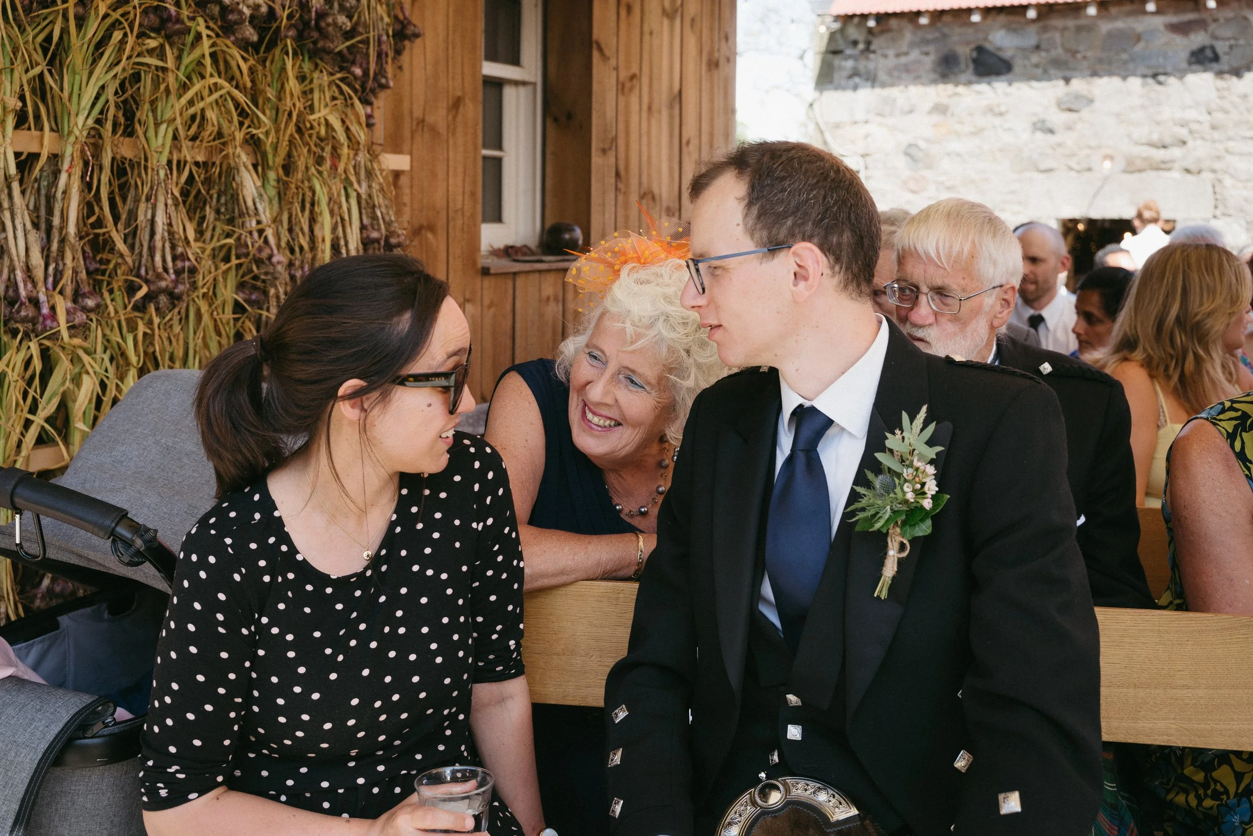 At a wedding reception, a woman in a polka dot dress and a man in a kilt are talking, with an elderly woman smiling between them, surrounded by seated guests in a rustic, wooden venue. - captured by an Edinburgh wedding photographer