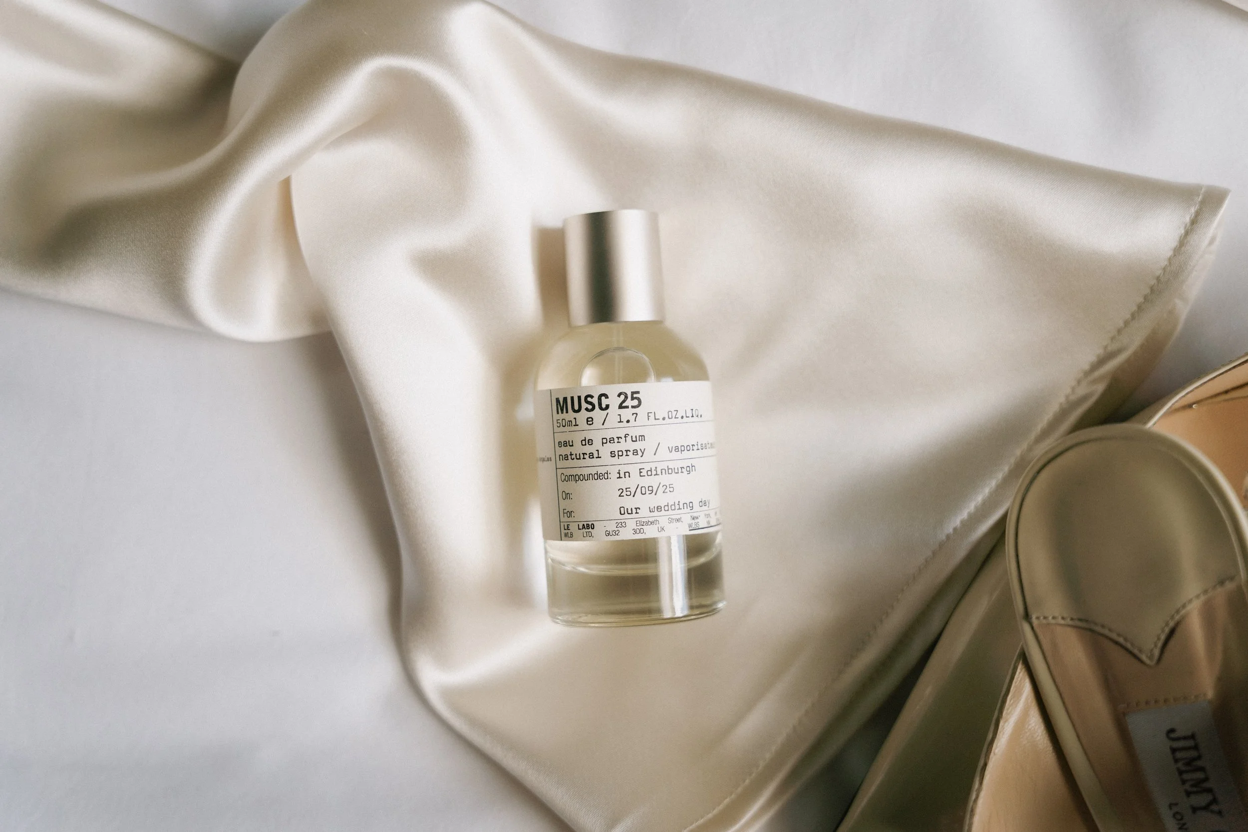 A bottle of perfume labeled 'MUSC 25' on a satin fabric surface with a pair of shiny beige shoes nearby. - captured by an Edinburgh wedding photographer