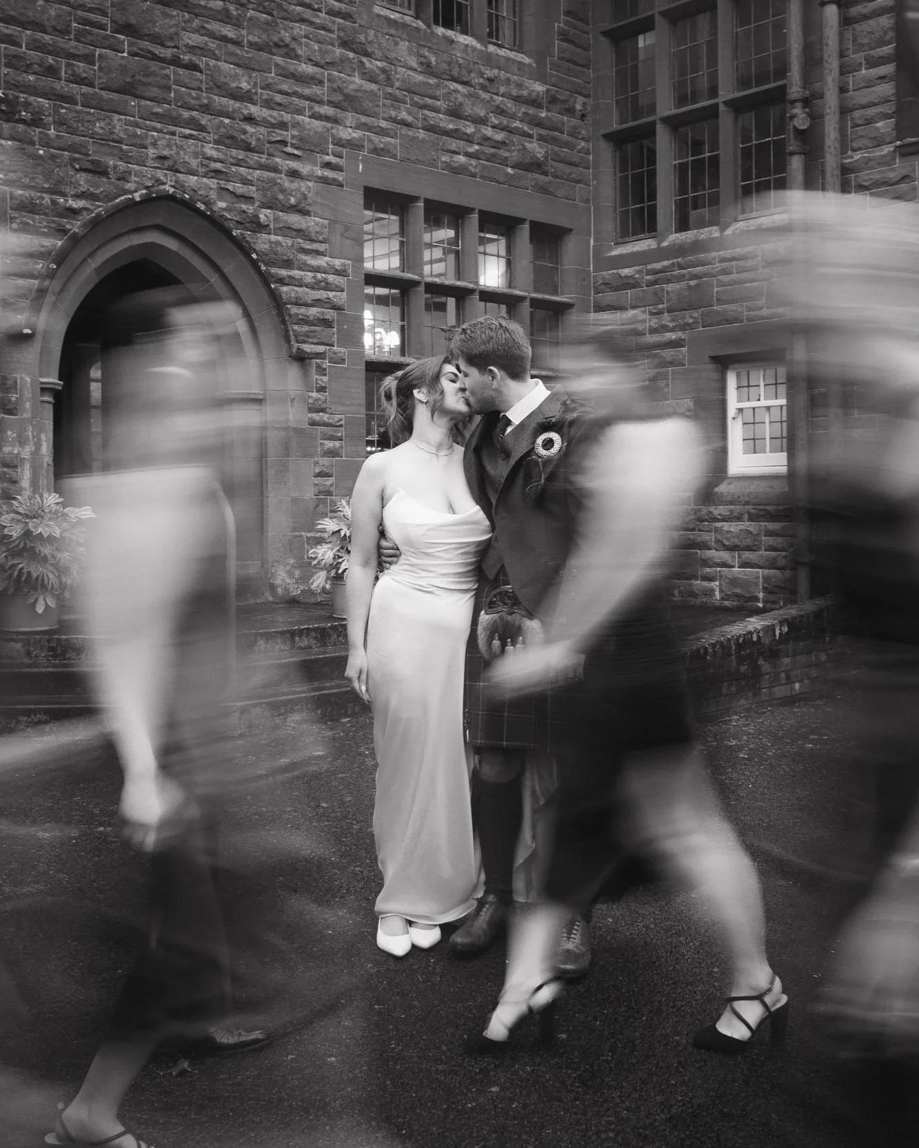 A wedding couple sharing a kiss outside a stone building with blurred figures walking by - captured by an Edinburgh wedding photographer