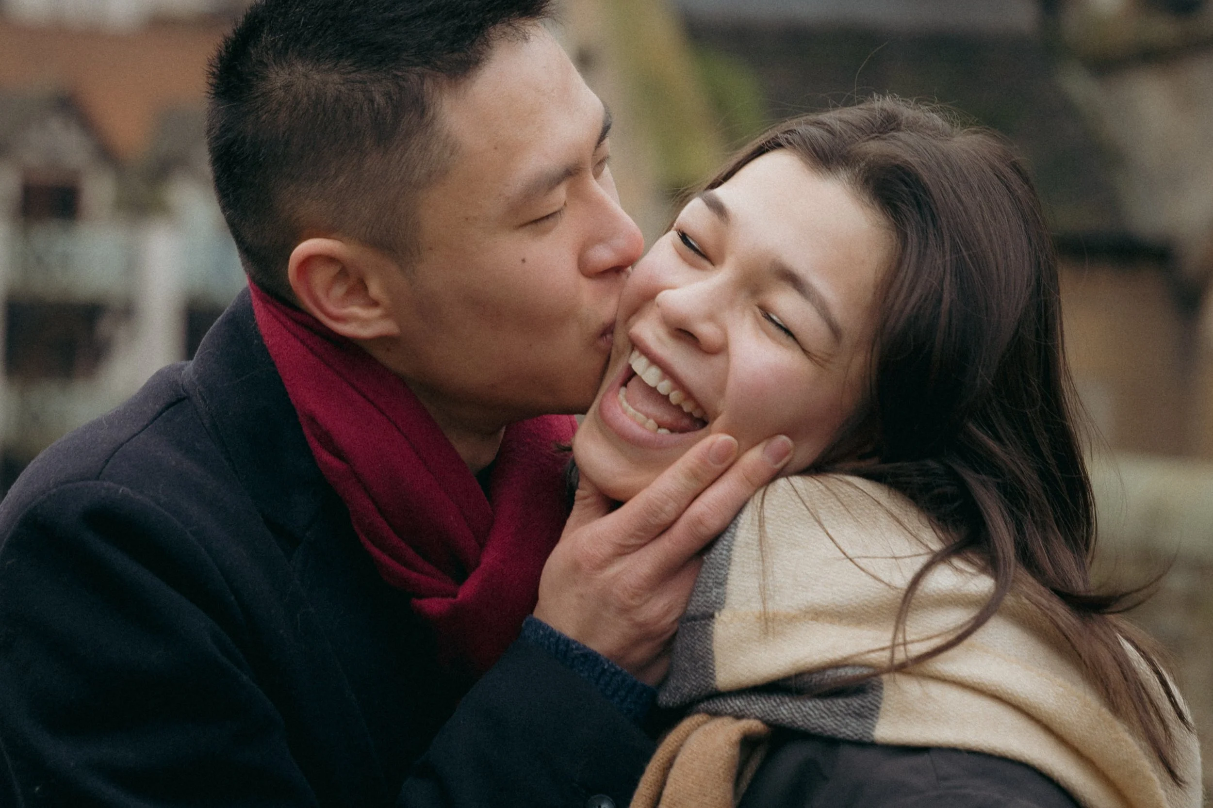 A man is kissing a woman on the cheek, and the woman is smiling and laughing, showing joy and affection. They appear to be outdoors, with a blurred background of buildings or houses. - captured by an Edinburgh wedding photographer