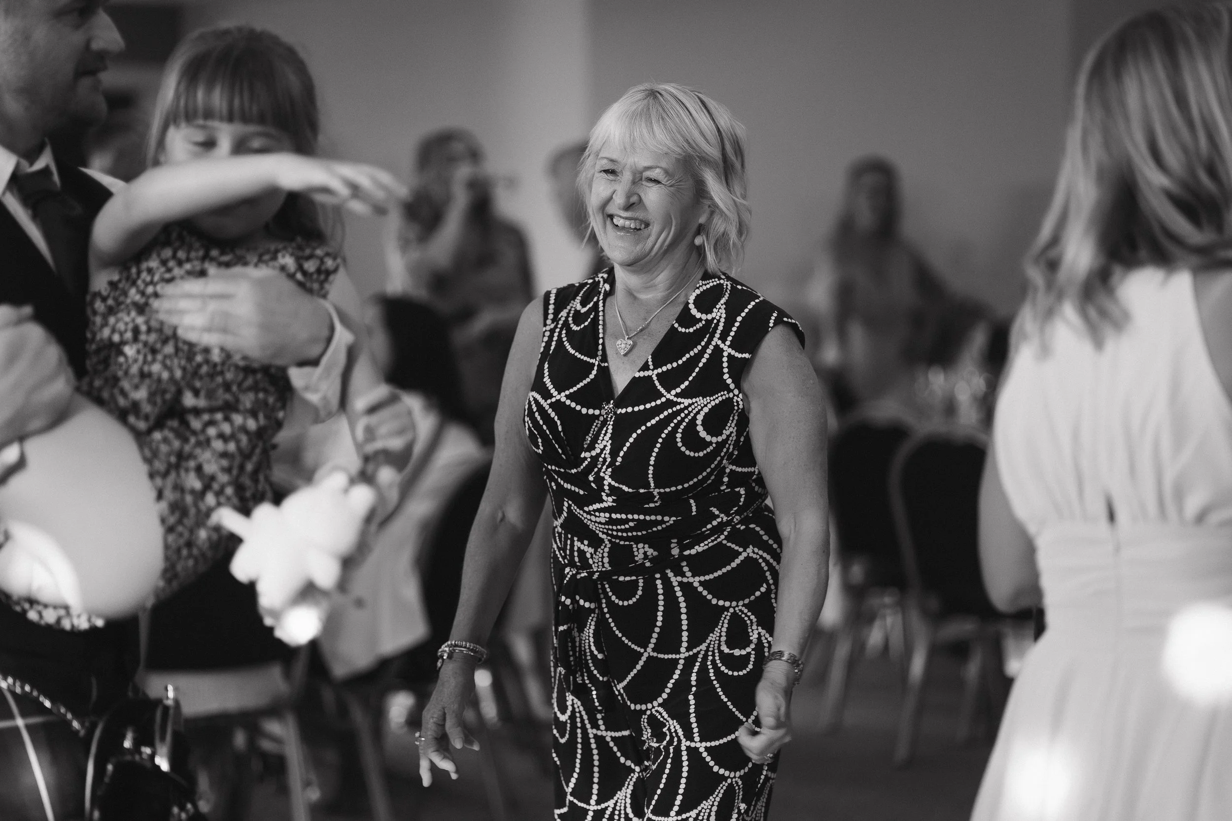 A woman smiling at an event, wearing a patterned dress, surrounded by people, some of whom are holding a child and taking photos. - captured by an Edinburgh wedding photographer