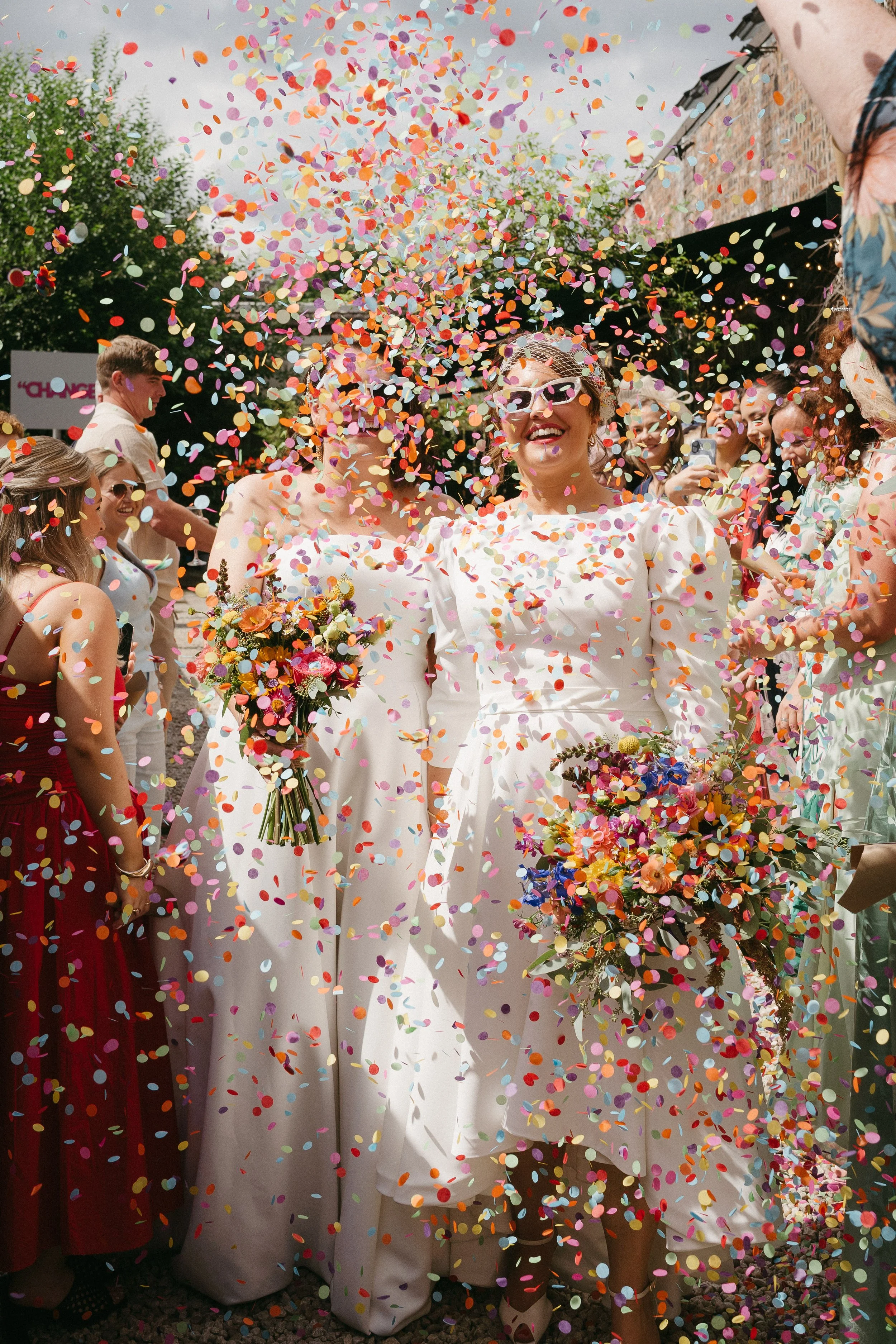 Two women dressed in white wedding dresses holding colorful flower bouquets, celebrating with multicolored confetti falling around them, surrounded by guests at an outdoor wedding - captured by an Edinburgh wedding photographer