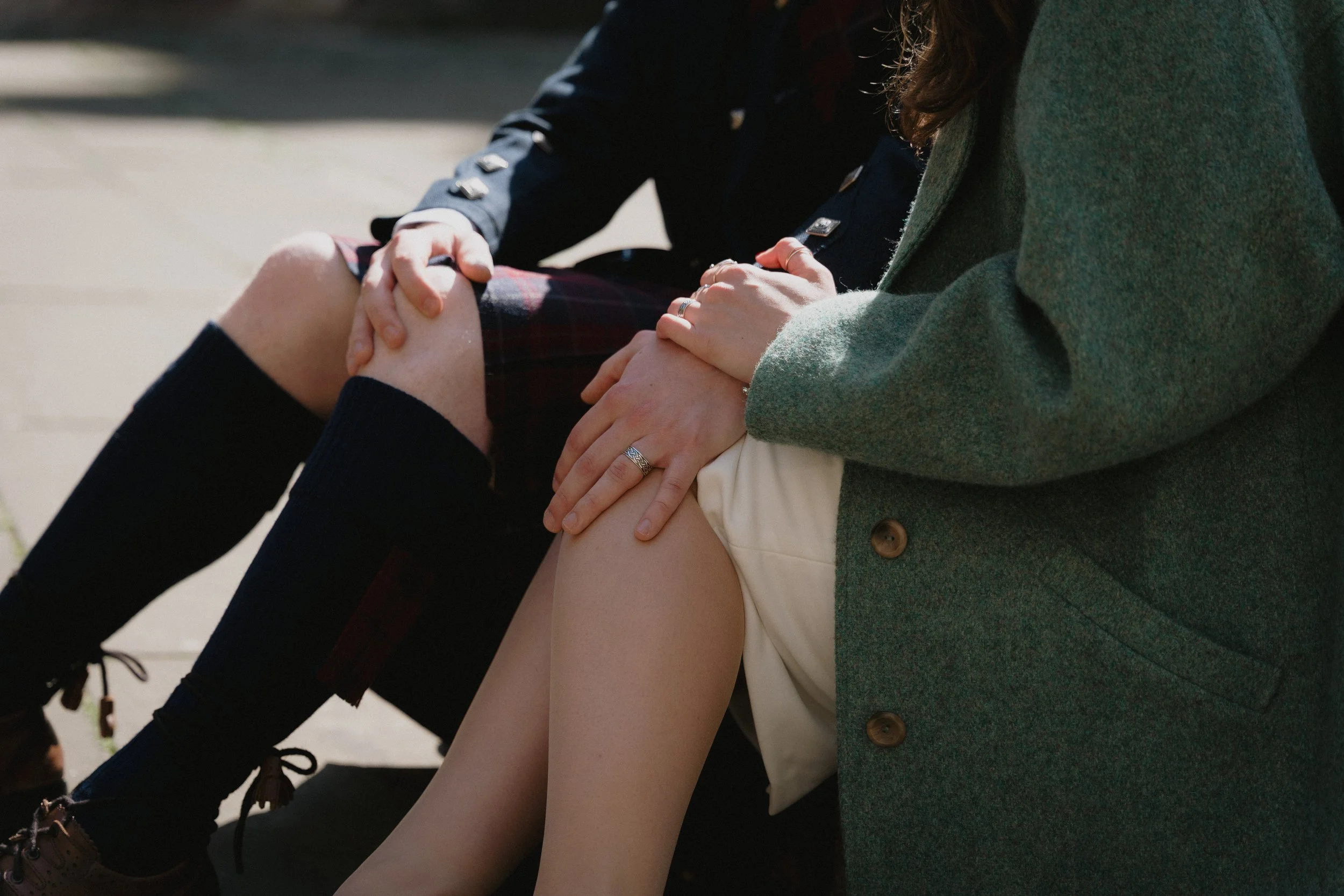 Two people sitting together outdoors, with one person holding the other's knee; the person on the left wears a navy blazer, tartan skirt, and knee-high socks, while the person on the right wears a green coat and beige skirt.