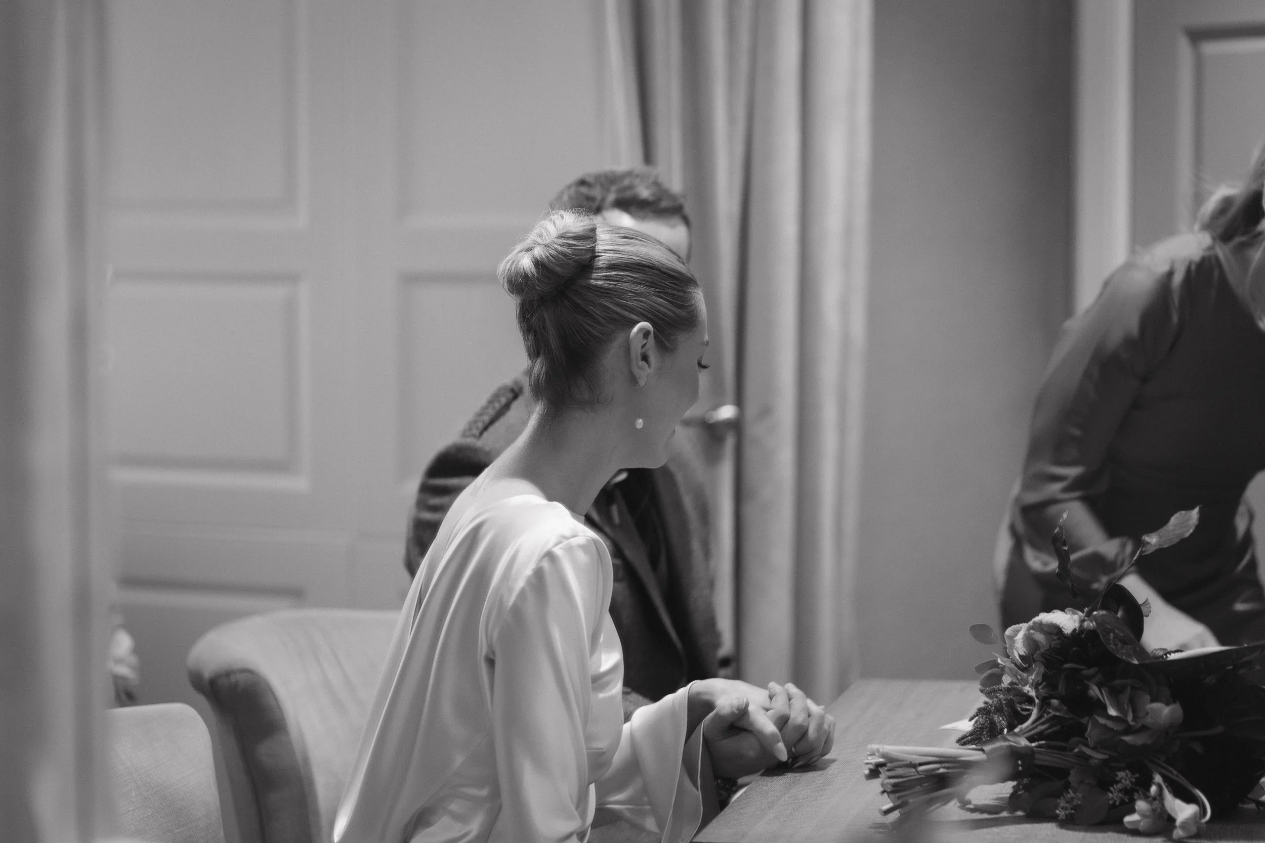 A woman with a bun hairstyle wearing a satin robe, sitting at a table with her hands clasped, with a bouquet of flowers in front of her, in a room with wood-paneled walls. - captured by an Edinburgh wedding photographer
