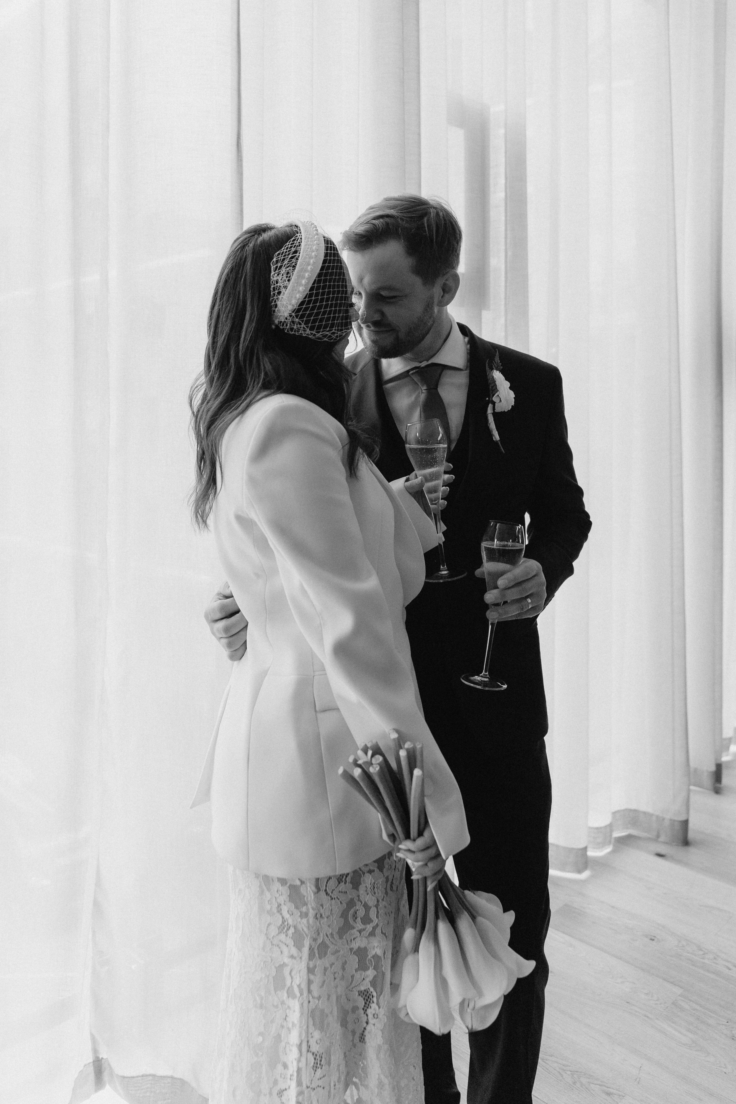 Black-and-white photo of a bride and groom sharing an intimate moment at their wedding, holding glasses of champagne, with the bride holding a bouquet. - captured by an Edinburgh wedding photographer