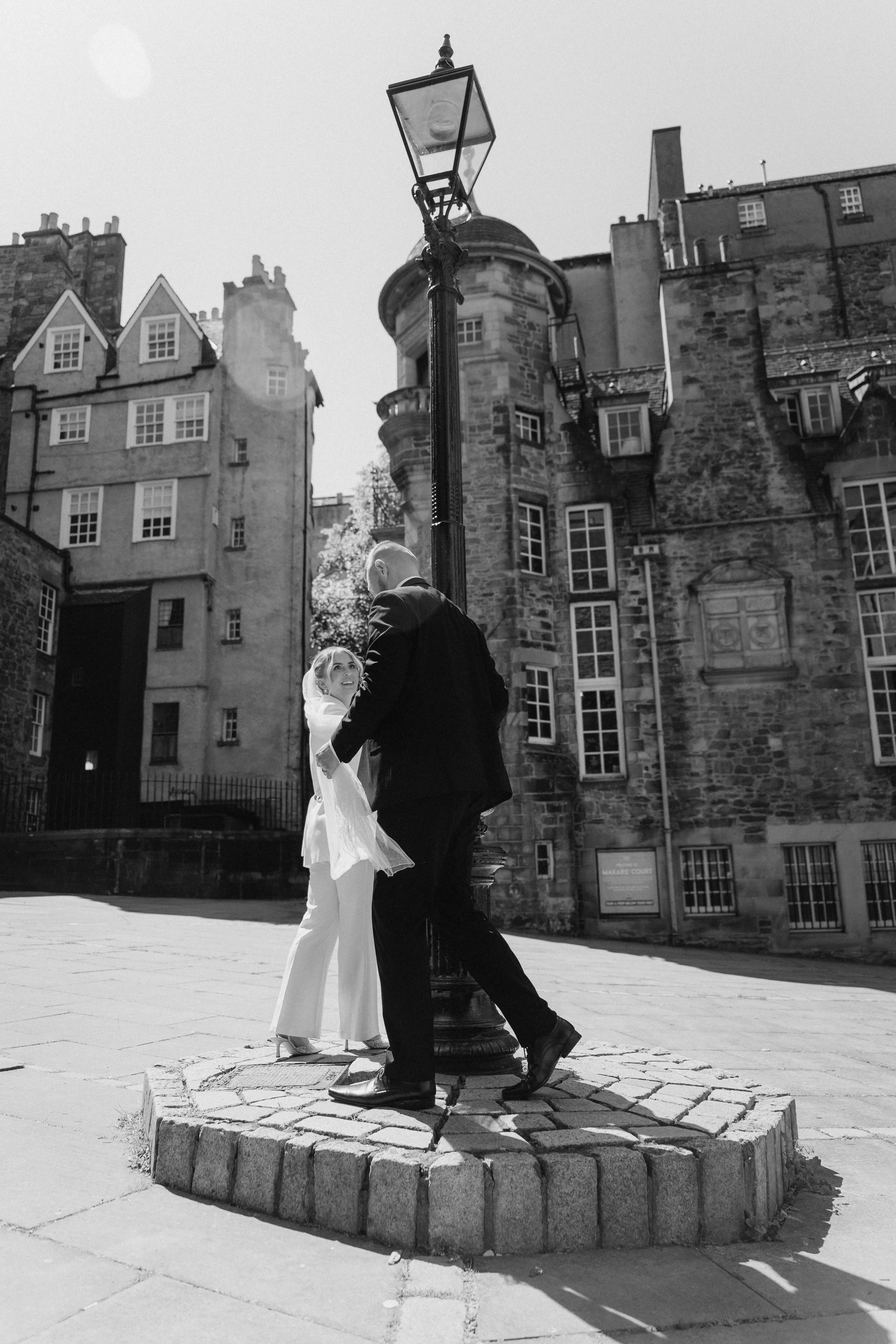 A couple in wedding attire standing close near a streetlamp in an urban setting with historic brick buildings in the background. - captured by an Edinburgh wedding photographer