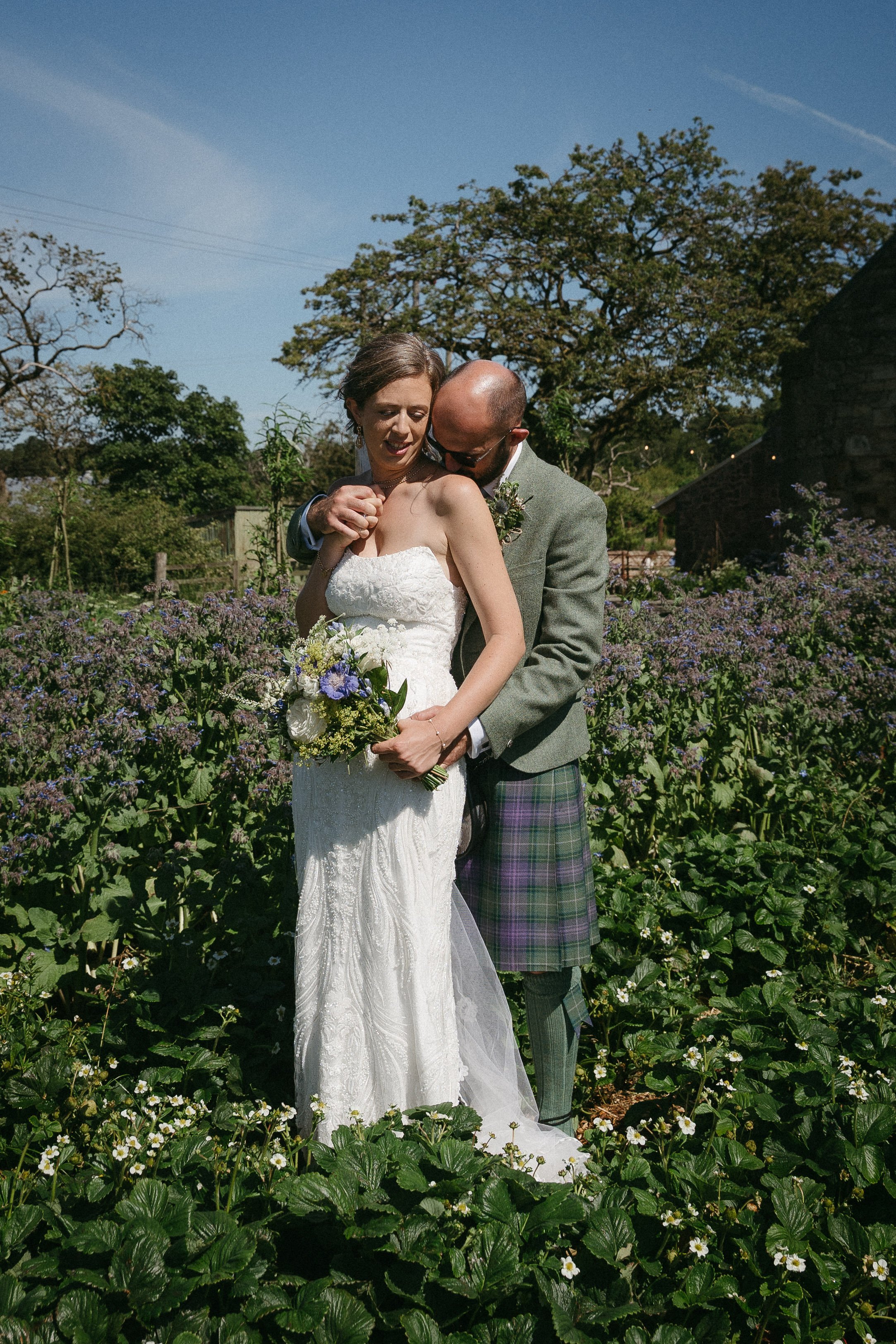 A bride and groom sharing an intimate moment in a lush garden with blooming flowers and large trees on a sunny day. - captured by an Edinburgh wedding photographer