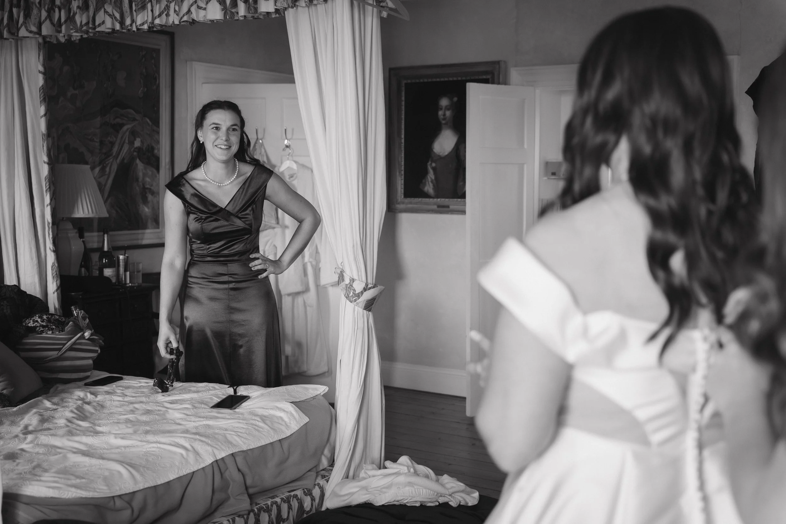 A woman in a satin dress talking to another woman who is in a white dress, with a necklace, in a room decorated with paintings and curtains. - captured by an Edinburgh wedding photographer