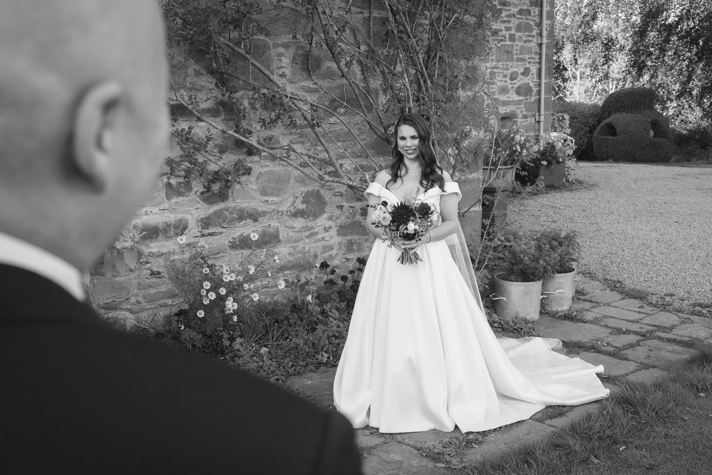 A bride in a white wedding gown holding a bouquet stands outdoors near a stone wall, smiling. A man, likely the groom, is visible in the foreground, out of focus. - captured by an Edinburgh wedding photographer