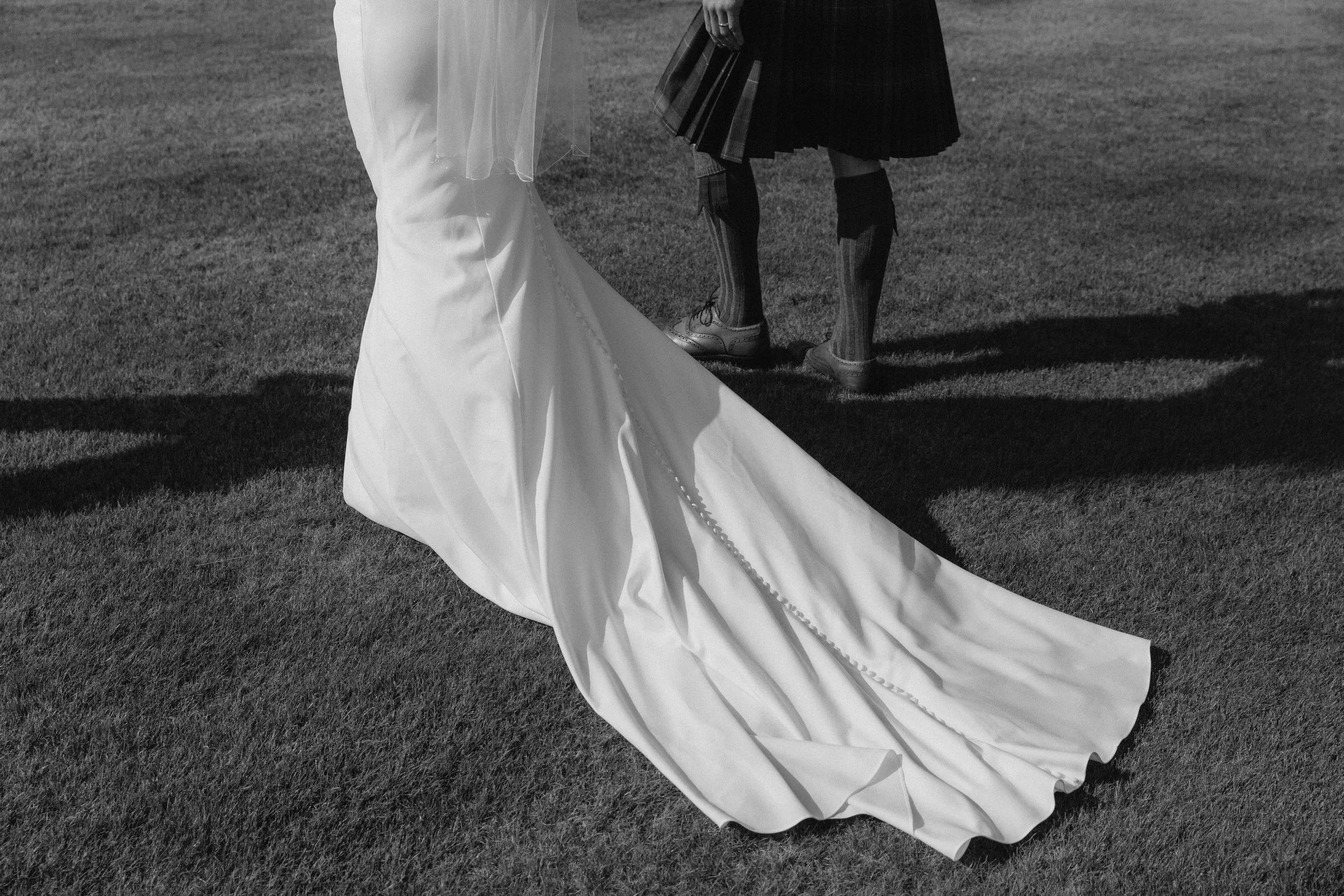 Black and white photo of a wedding scene with a bride in a long white dress and train, and a person in traditional Scottish attire including a kilt and laced shoes, standing on grass. - captured by an Edinburgh wedding photographer