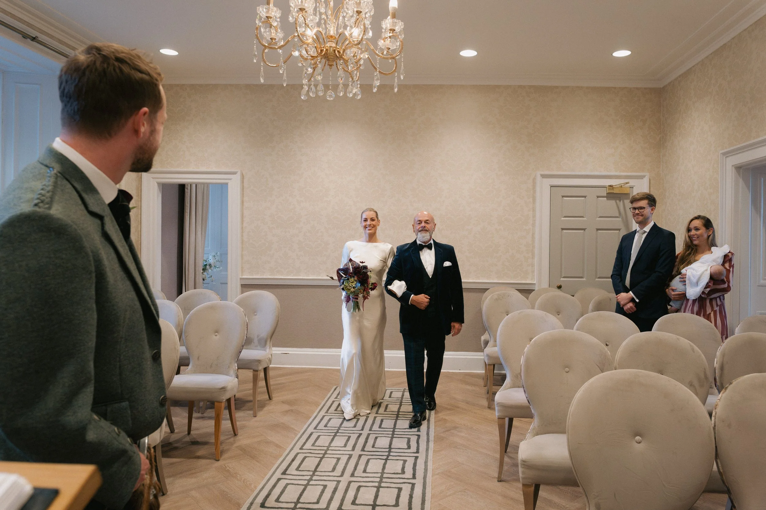 Bride walking down the aisle with her father in a wedding ceremony inside a decorated room with cream-colored walls, chandelier, and empty chairs. - captured by an Edinburgh wedding photographer