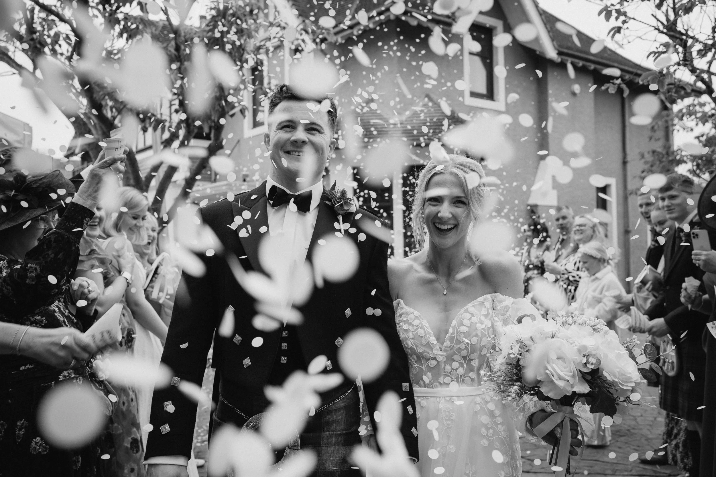 A newly married couple walking through a confetti celebration outside, surrounded by smiling guests, with a house in the background, in black and white - captured by an Edinburgh wedding photographer