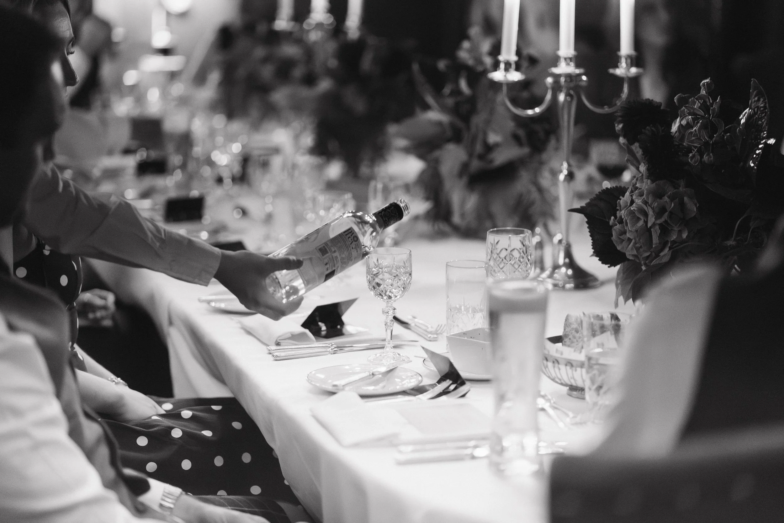A person pouring water into a decorative glass on a formal dining table with elegant glassware, a candelabra, and floral centerpieces, in black and white. - captured by an Edinburgh wedding photographer