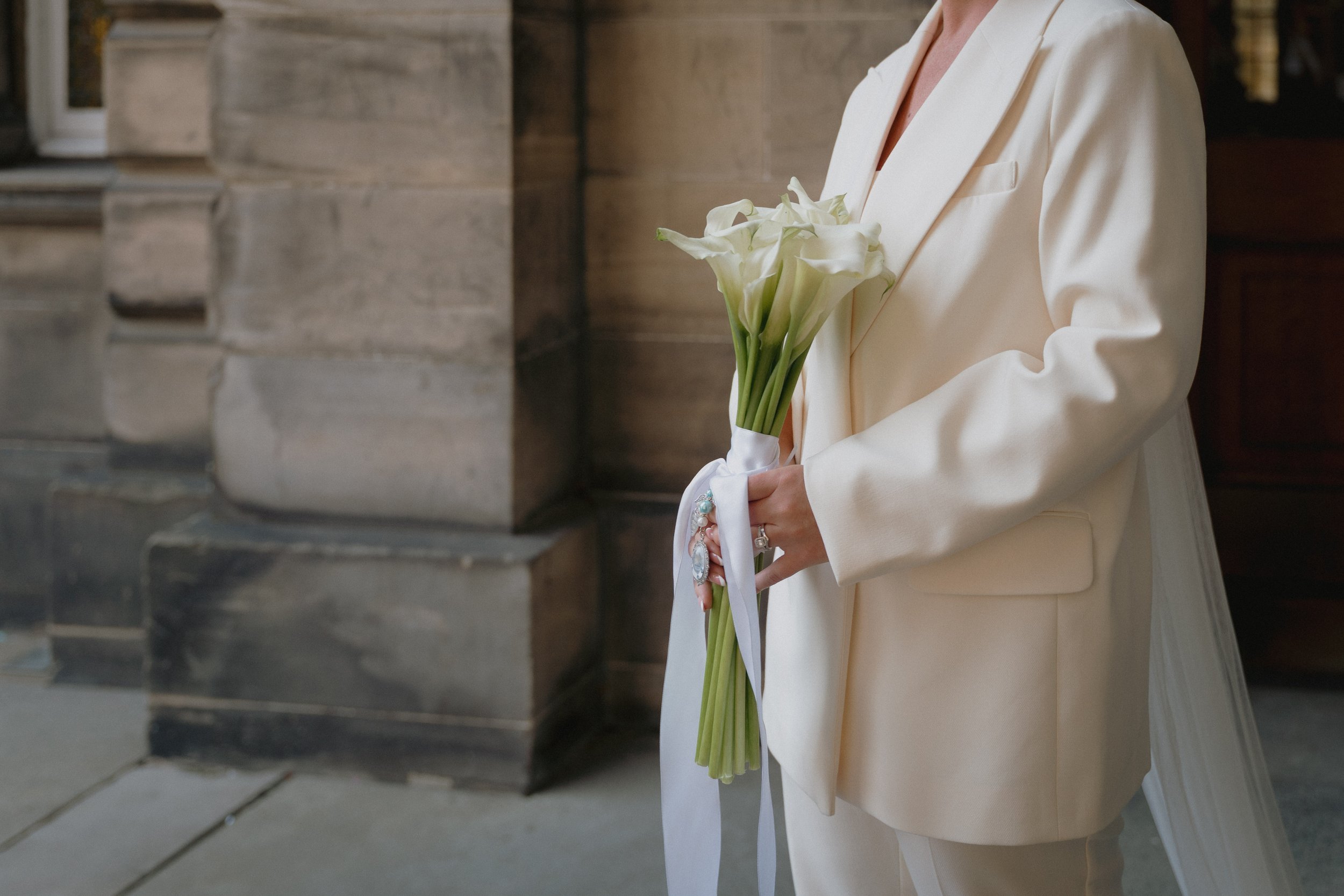 A person dressed in a cream-colored suit holding a bouquet of white calla lilies wrapped with a white ribbon. - captured by an Edinburgh wedding photographer