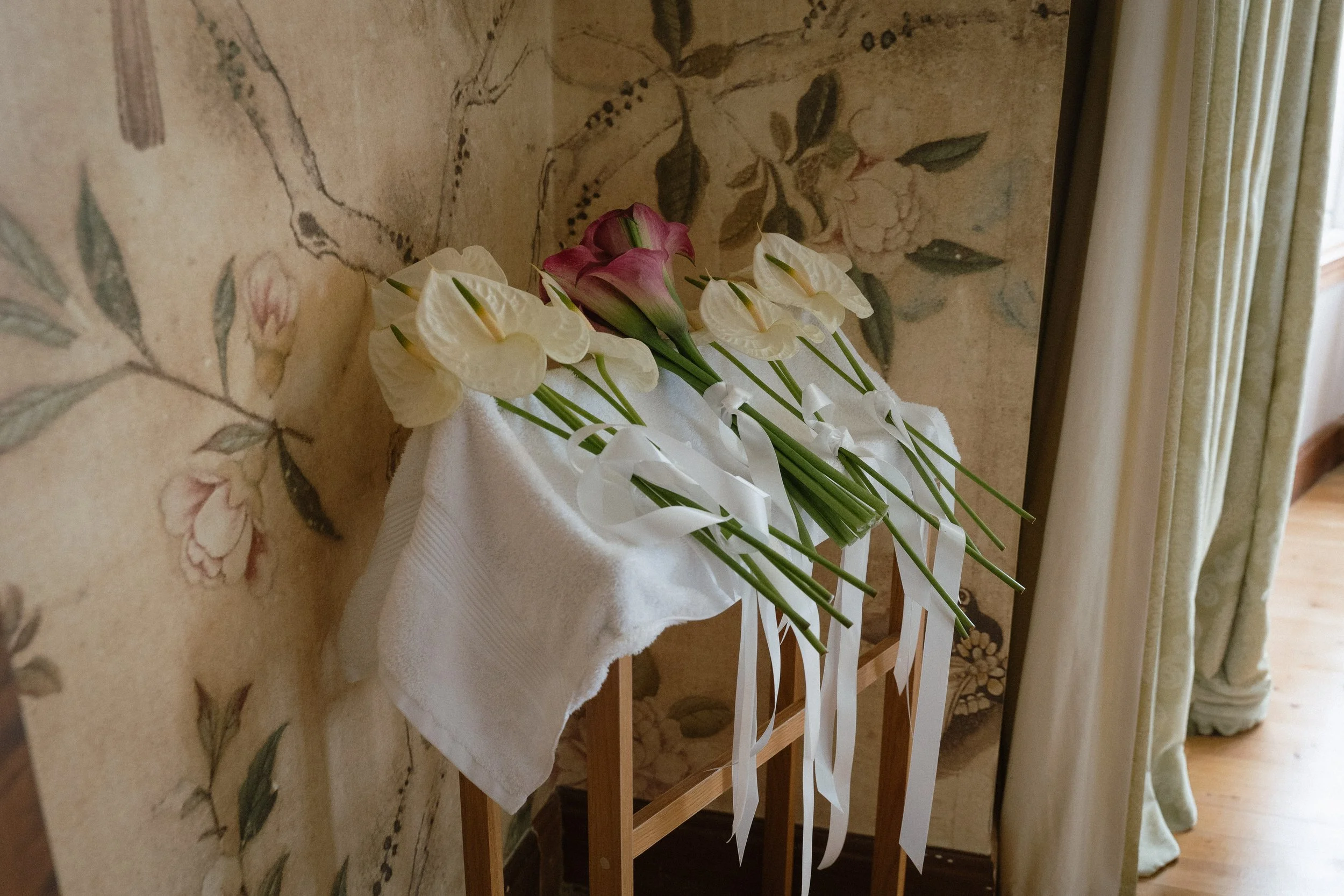 A bouquet of white and pink calla lilies with long green stems and white ribbons, resting on a white cloth draped over a wooden stand, against a floral wallpaper background. - captured by an Edinburgh wedding photographer