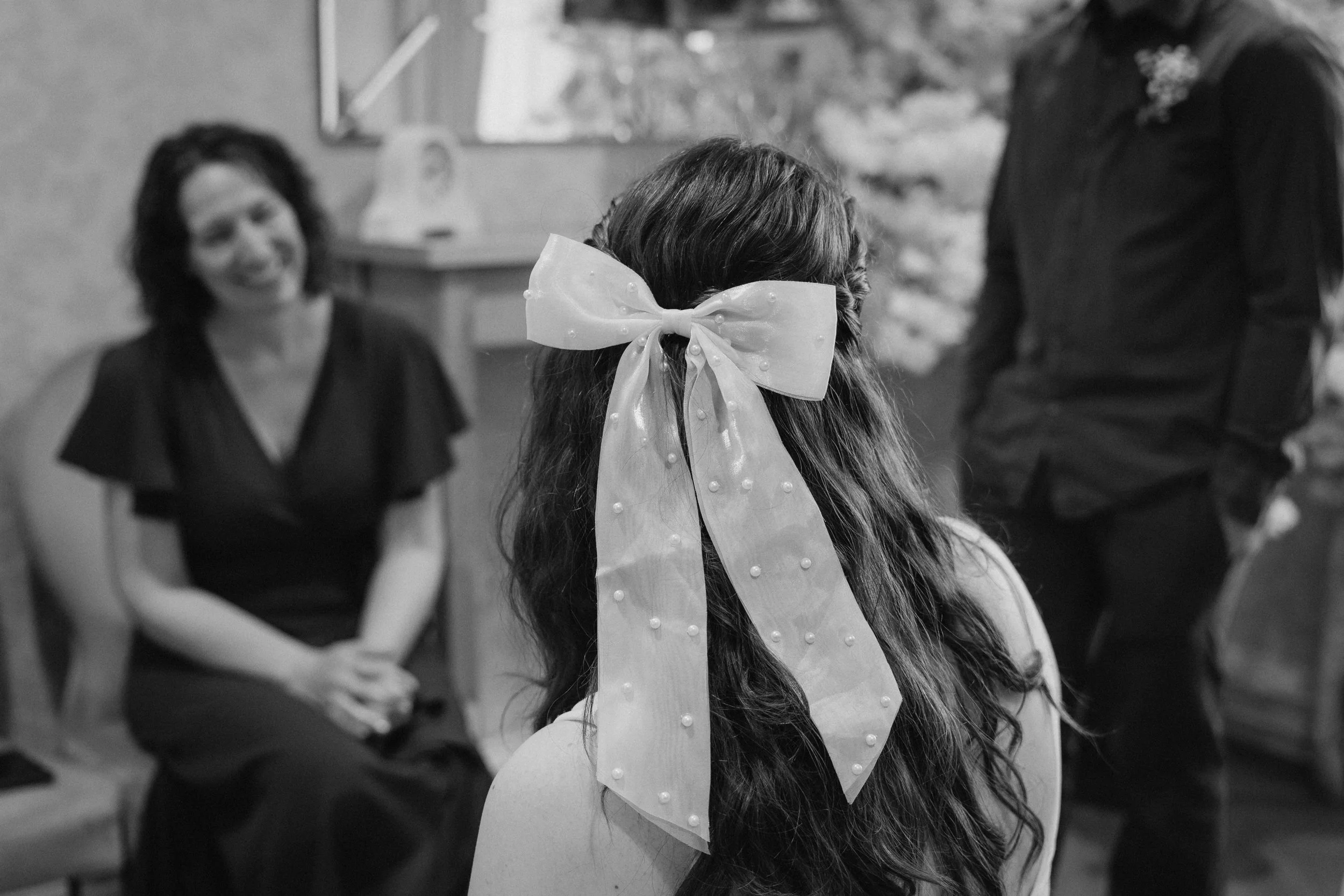 A young girl with long hair wearing a large bow on her head, sitting with her back to the camera, in a room with four adults, one woman smiling in the background and others standing or sitting nearby. - captured by an Edinburgh wedding photographer