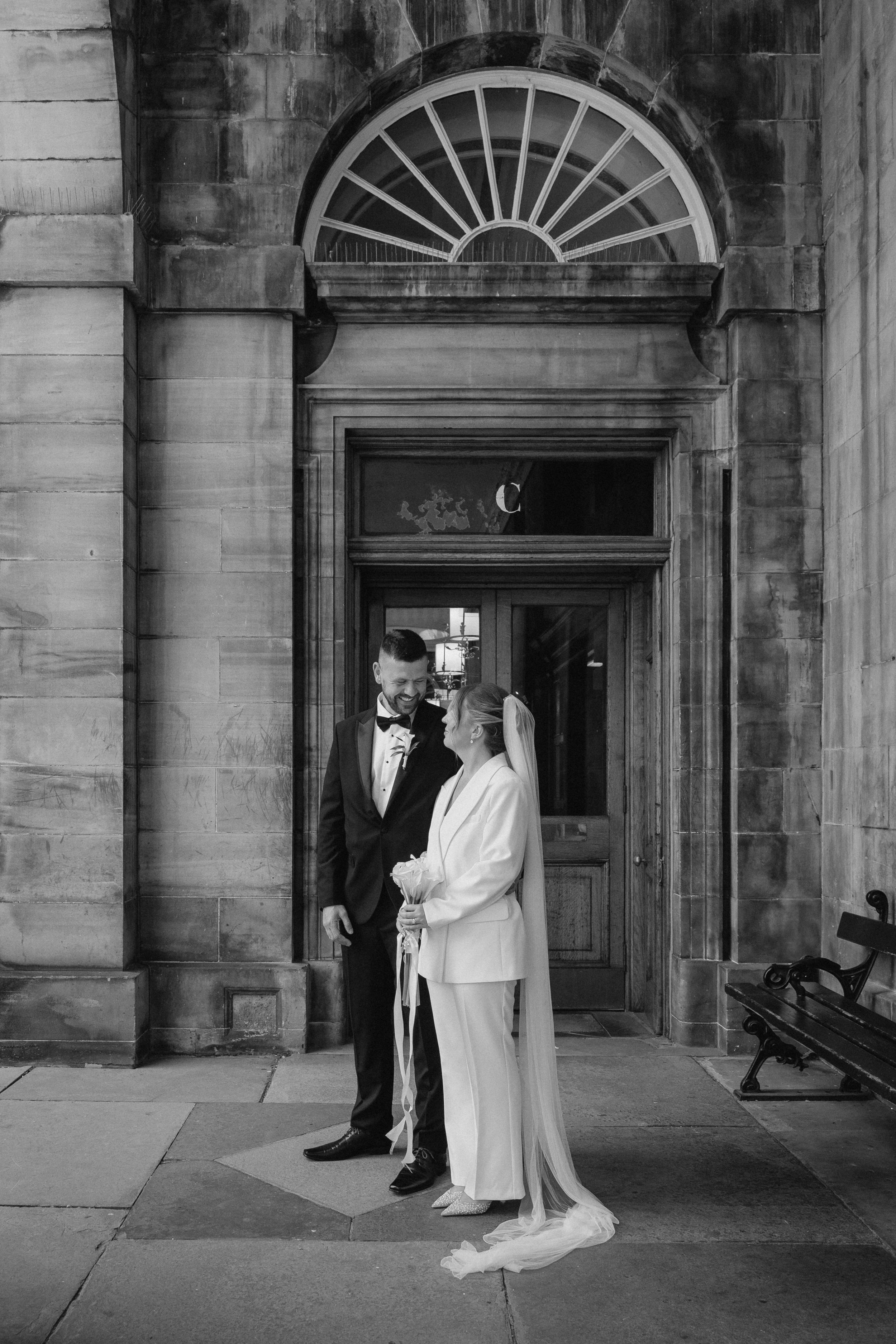 A couple, dressed in wedding attire, standing outside a building, smiling and looking at each other. The woman is holding a bouquet and wearing a bridal veil, while the man is in a tuxedo.