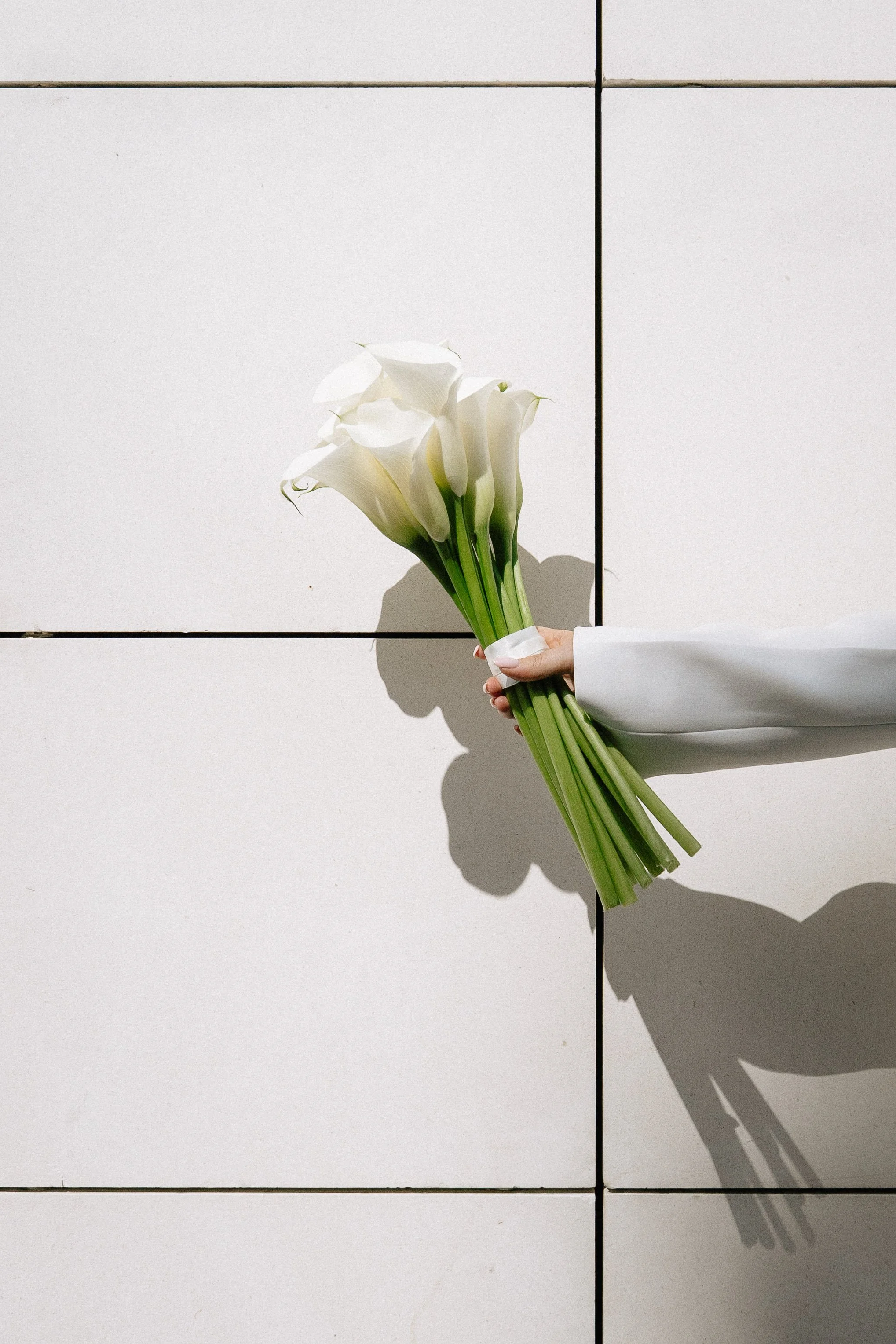 Person holding a bouquet of white calla lilies against a white tiled wall, with shadows cast by the flowers. - captured by an Edinburgh wedding photographer