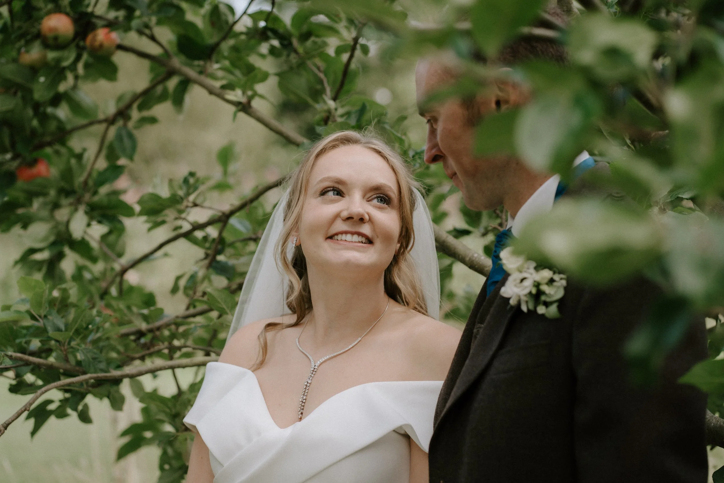 A bride and groom share a moment under a tree during their wedding, smiling at each other, surrounded by green leaves. - captured by an Edinburgh wedding photographer