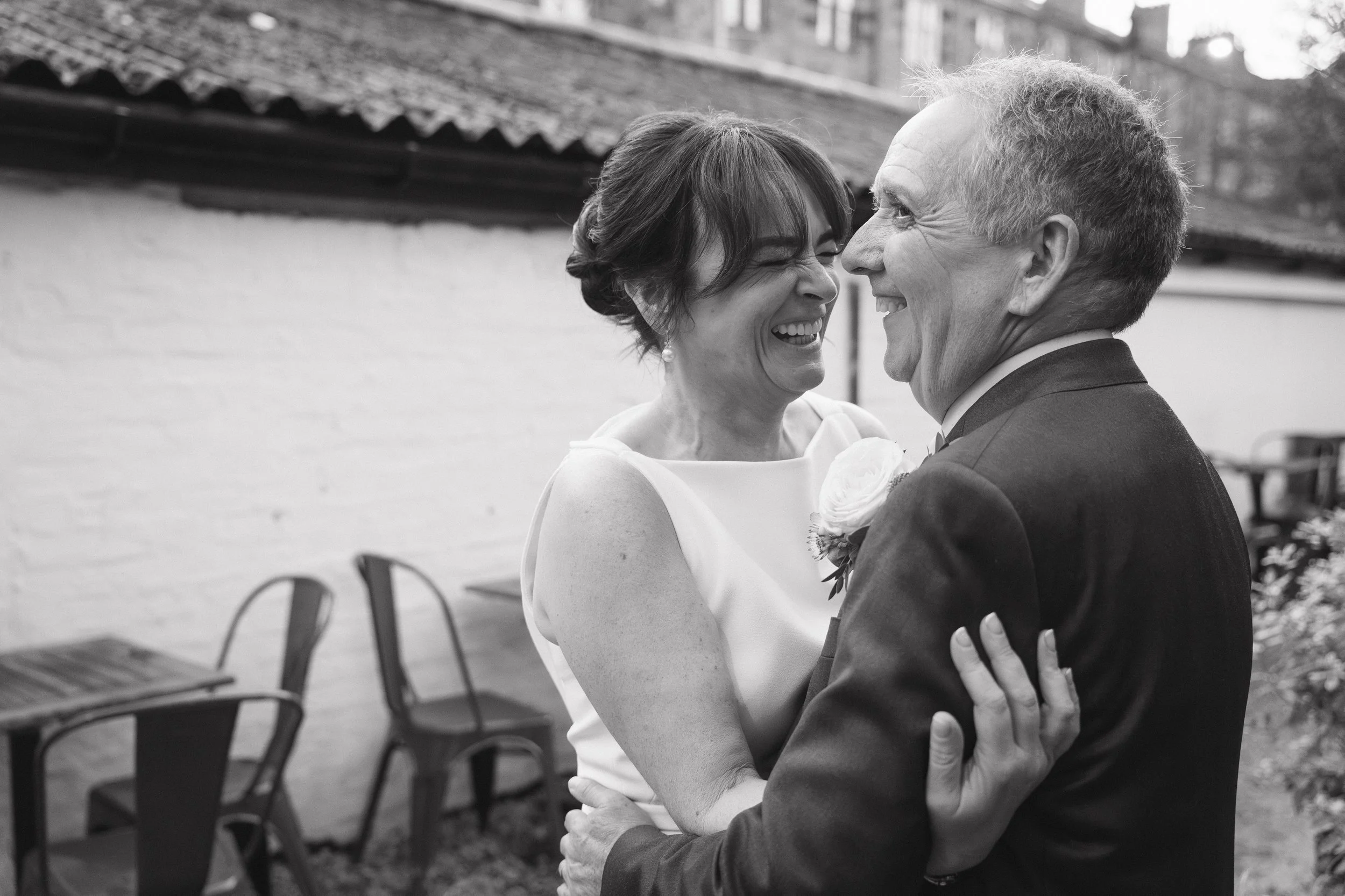 A black-and-white photo of a couple, likely at a wedding, sharing a joyful moment as they hold each other. The woman is smiling widely with her eyes closed, and the man is smiling at her, holding her arm. They are outdoors, with a brick wall, fence, 